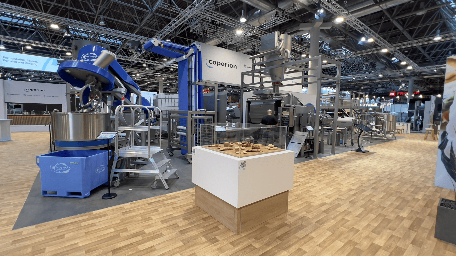 Ein Messestand präsentiert große industrielle Lebensmittelverarbeitungsanlagen der Marke Coperion. Ein weißer Sockel davor präsentiert Muster kleiner Gebäcke. Der Stand befindet sich in einer geräumigen Halle mit Holzboden.