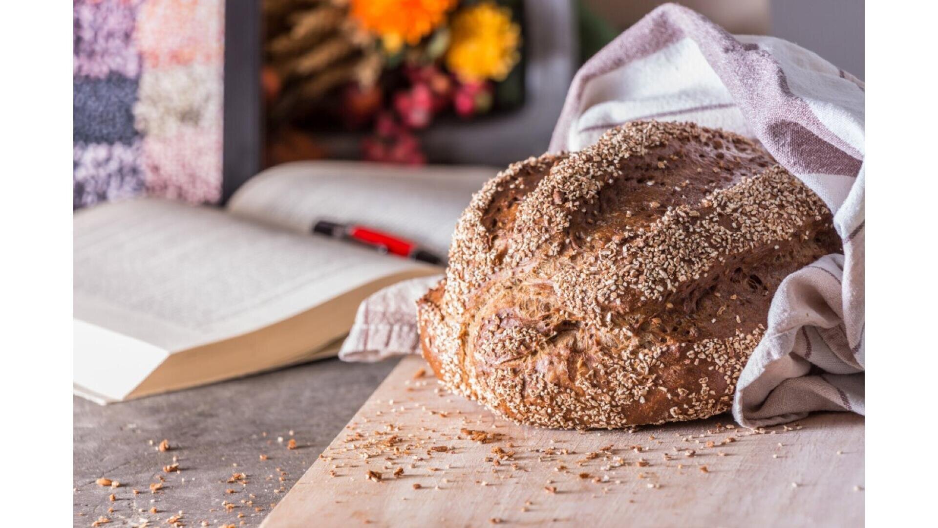 Ein Laib Körnerbrot liegt auf einem Holzbrett, teilweise in ein gestreiftes Tuch gewickelt. Ein aufgeschlagenes Buch, ein roter Stift und eine Vase mit bunten Blumen sind unscharf im Hintergrund zu erkennen.