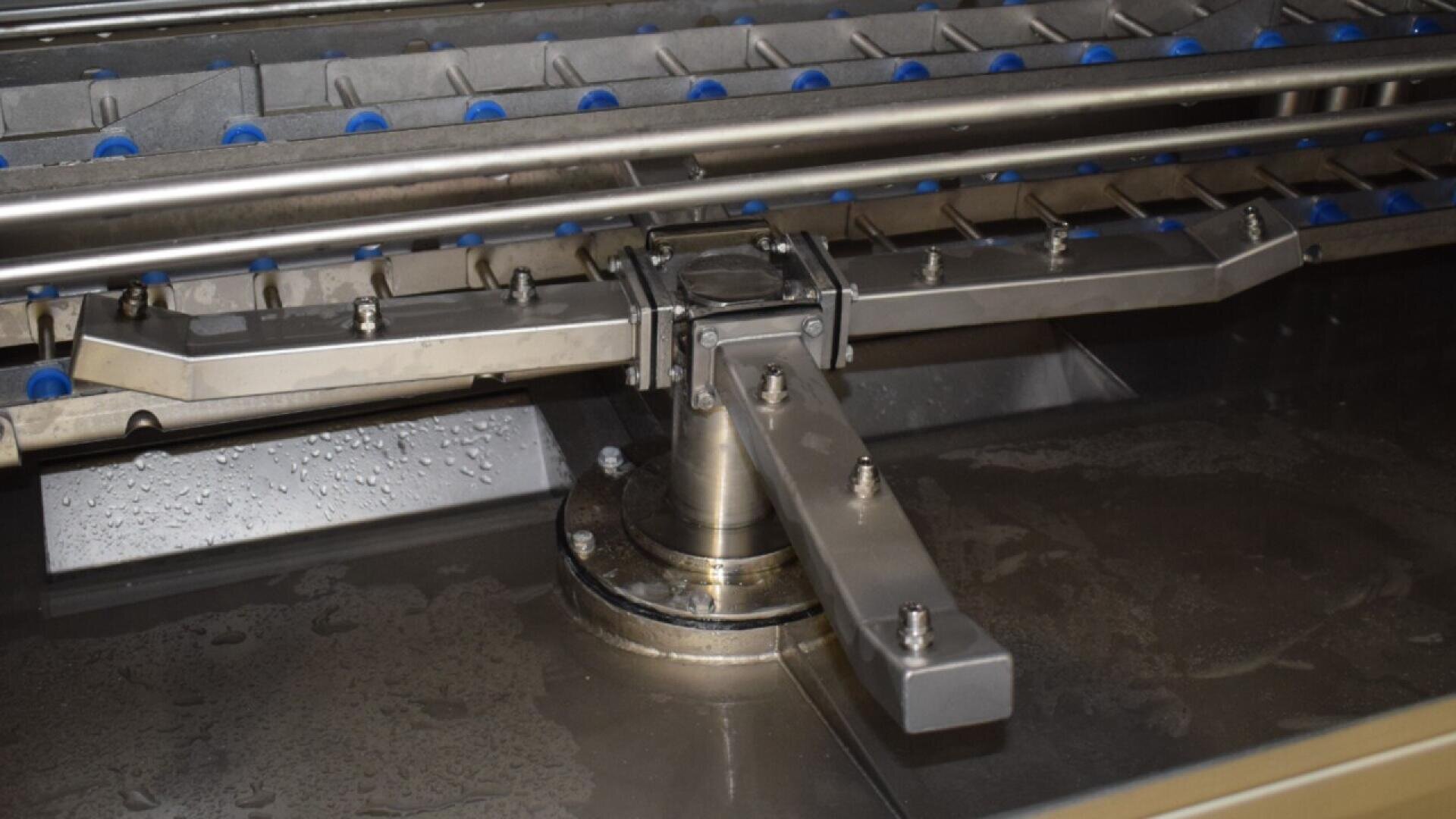 A close-up view of an industrial machine with metal arms, rails, bolts, and blue rollers, possibly used for processing or transporting items. The surrounding surface has water droplets and wet areas.