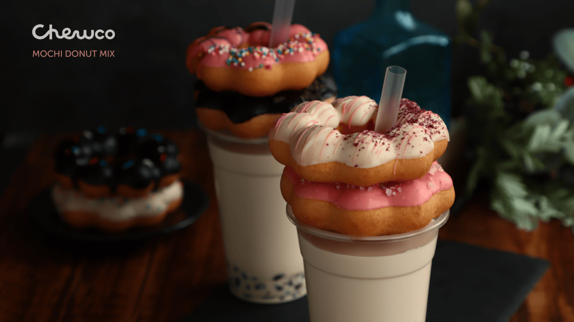 Two cups of bubble tea, each topped with two colorful mochi donuts, sit on a dark surface. The donuts are decorated with pink, white, and chocolate glazes and sprinkles. Blurred greenery and another donut are in the background.