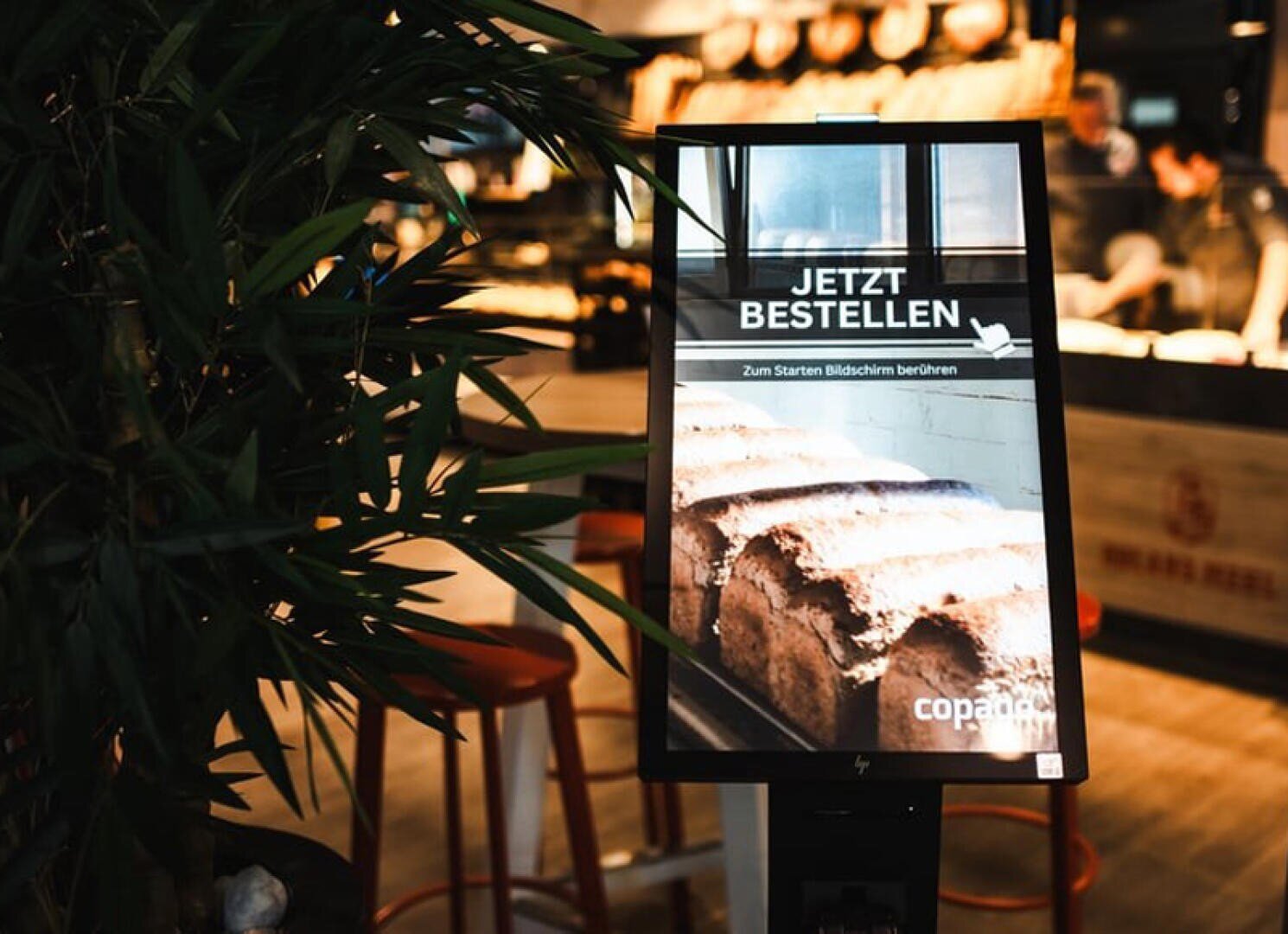 Auf dem Bildschirm eines digitalen Bestellkiosks steht auf Deutsch „Jetzt bestellen“ und ein Bild von Brot in einer Bäckerei, umgeben von Pflanzen und Hockern in einem warm beleuchteten Laden.