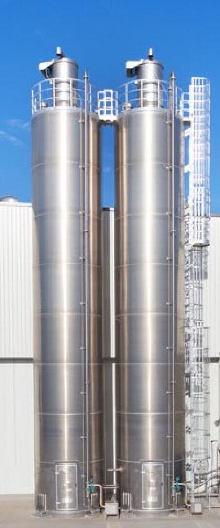 Two tall, shiny stainless steel silos stand side by side against a white industrial building, with a metal ladder and platform structure on the right side and a clear blue sky in the background.