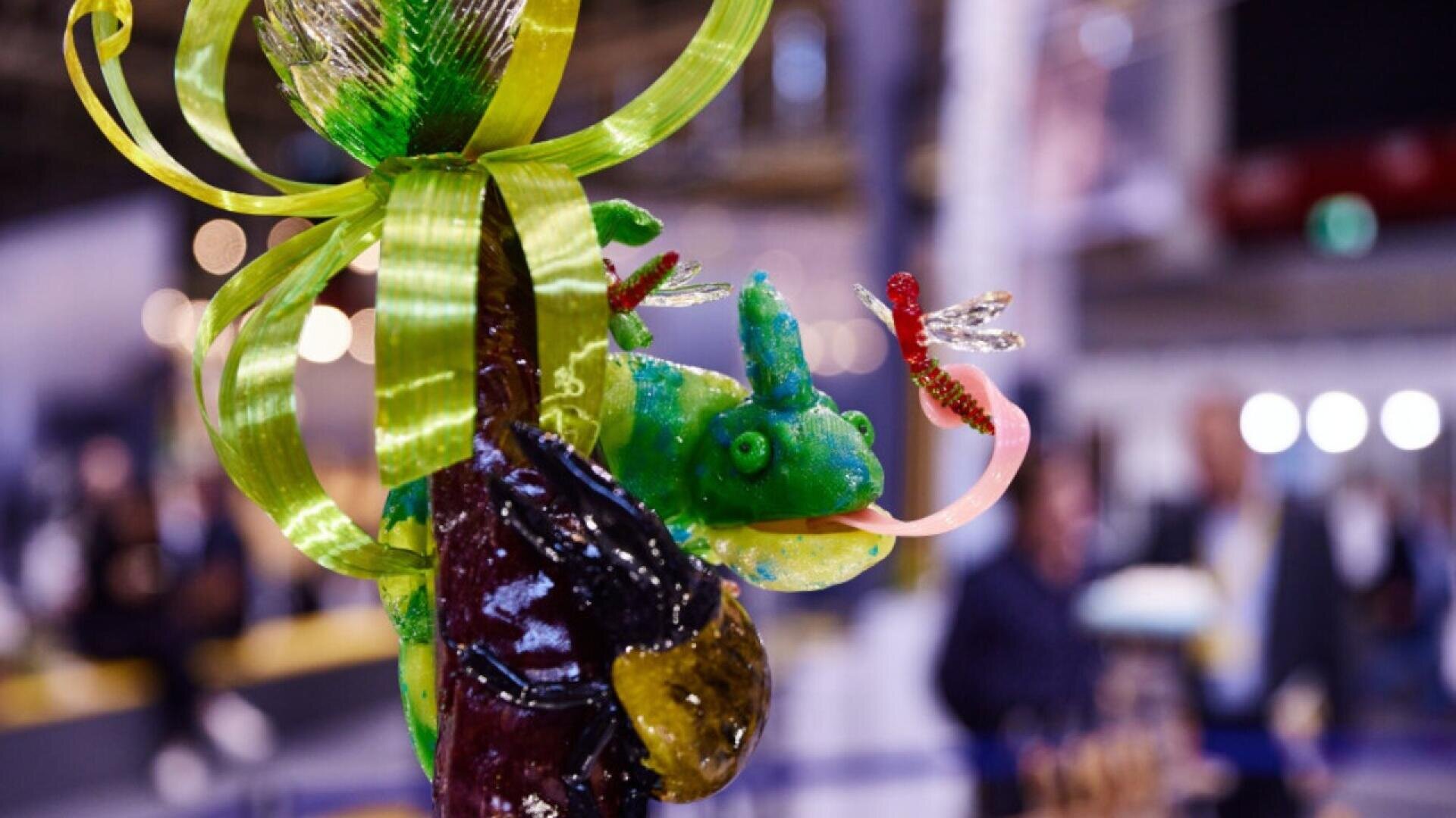 A colorful sugar sculpture of a green chameleon with a long pink tongue and two dragonflies, decorated with a shiny yellow ribbon, displayed in an indoor setting with blurred people in the background.