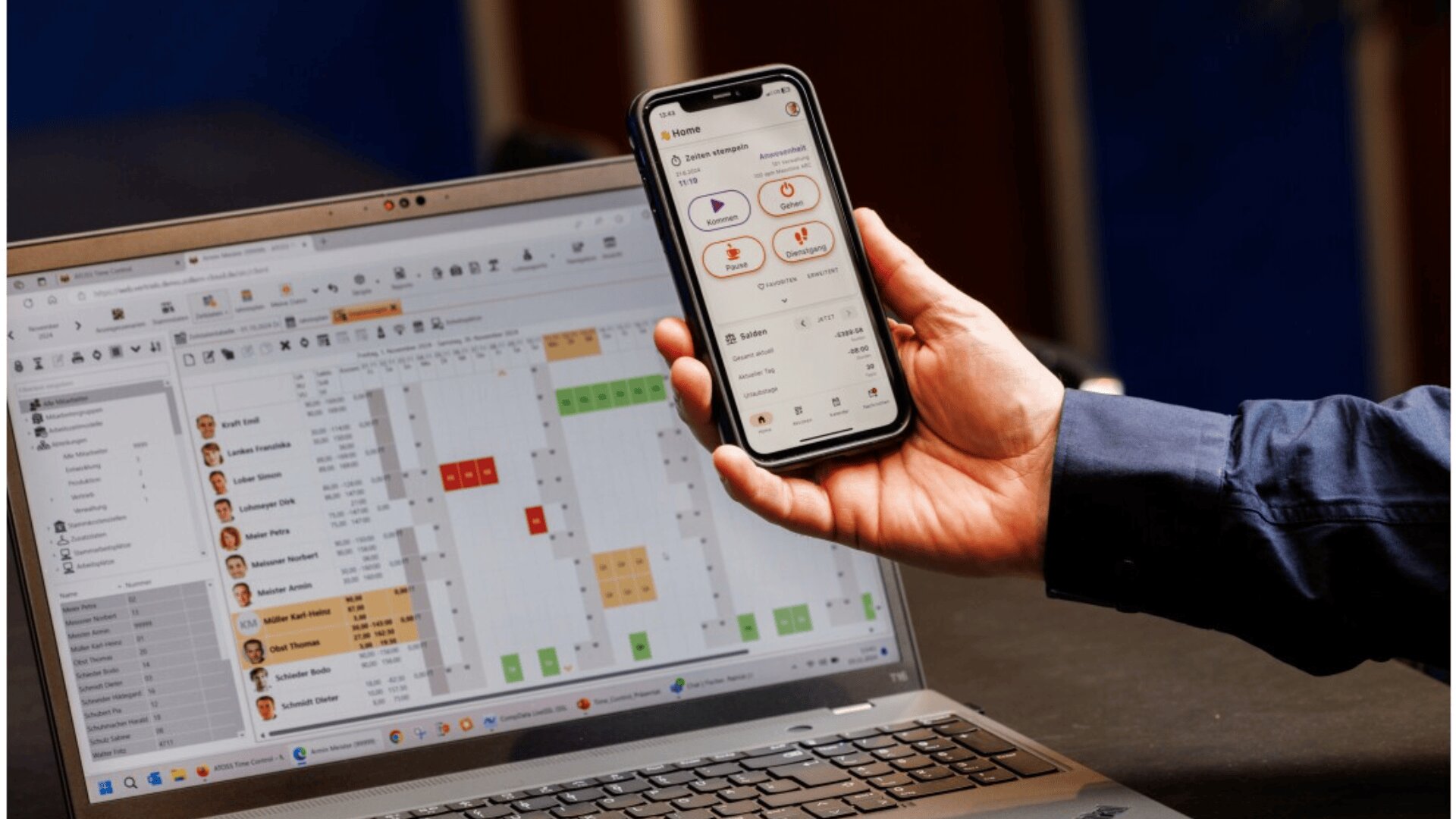 A person holds a smartphone displaying an app with icons and graphs, while a laptop nearby shows a scheduling or calendar application with names, dates, and color-coded events.