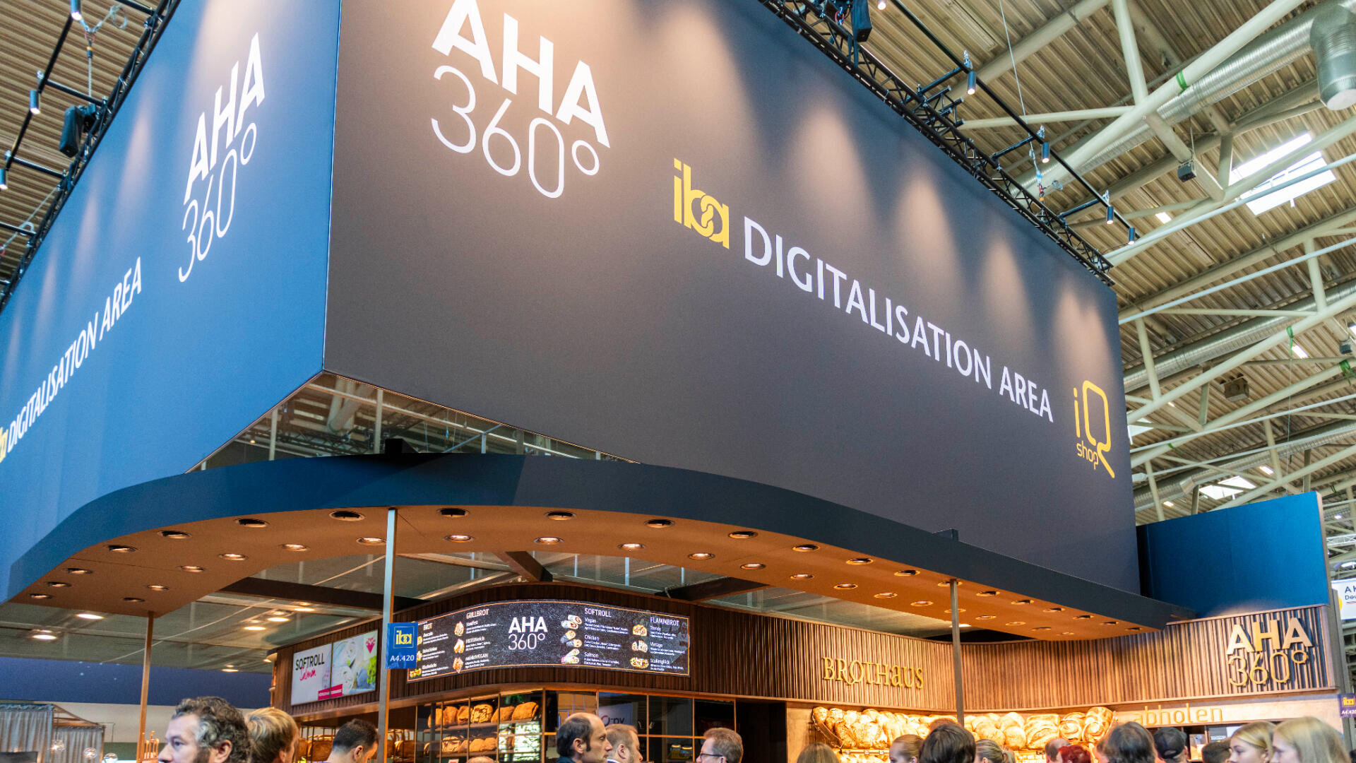 Großer Messestand mit den Aufschriften „AHA 360°“ und „DIGITALISATION AREA“ auf den Deckenschildern, in einem hellen, modernen Ambiente, mit Menschen, die sich darunter versammelt haben, und Regalen, in denen verschiedene Brotsorten ausgestellt sind.