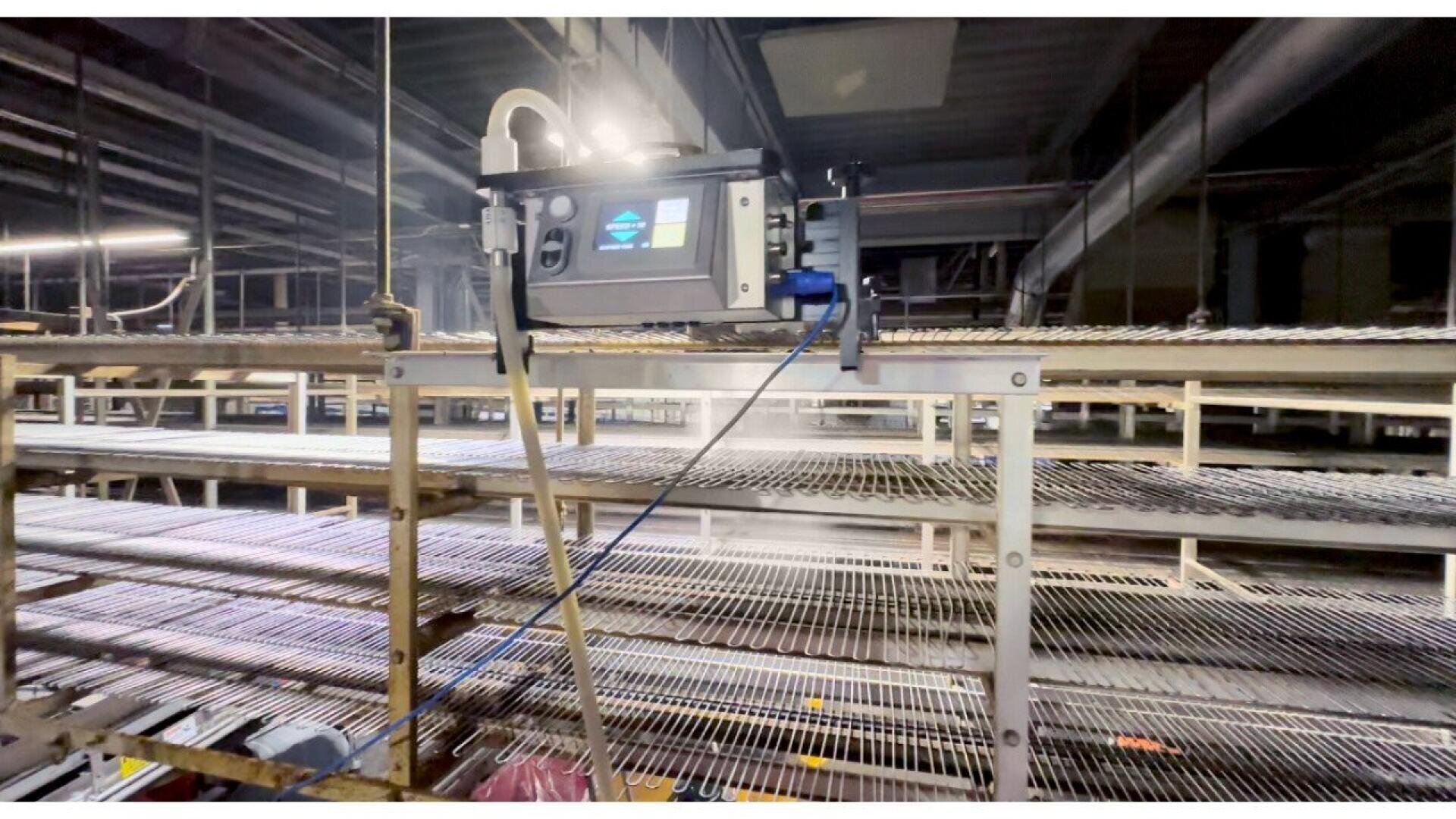 Ein industrielles Gerät, das in einer Fabrik über Metallregalen angebracht ist und Nebel oder Dampf über mehrere horizontale Regale abgibt, möglicherweise zur Desinfektion oder Feuchtigkeitsregelung. Der Bereich ist mit Oberlicht beleuchtet.