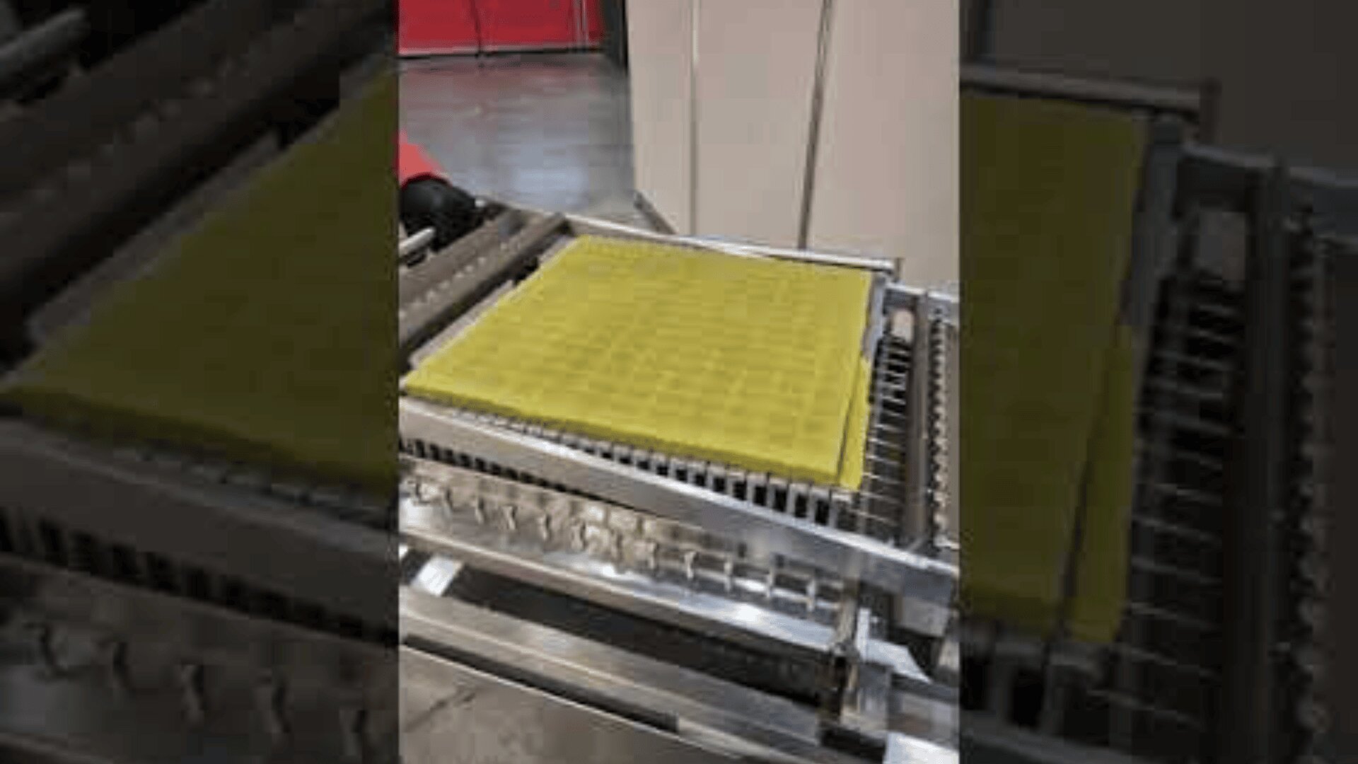 Ein Metallförderer hält ein gelbes, rechteckiges Tablett mit einem Gittermuster, das möglicherweise in einem Lager oder einer Fabrik für die Lebensmittel- oder Industrieverarbeitung verwendet wird.