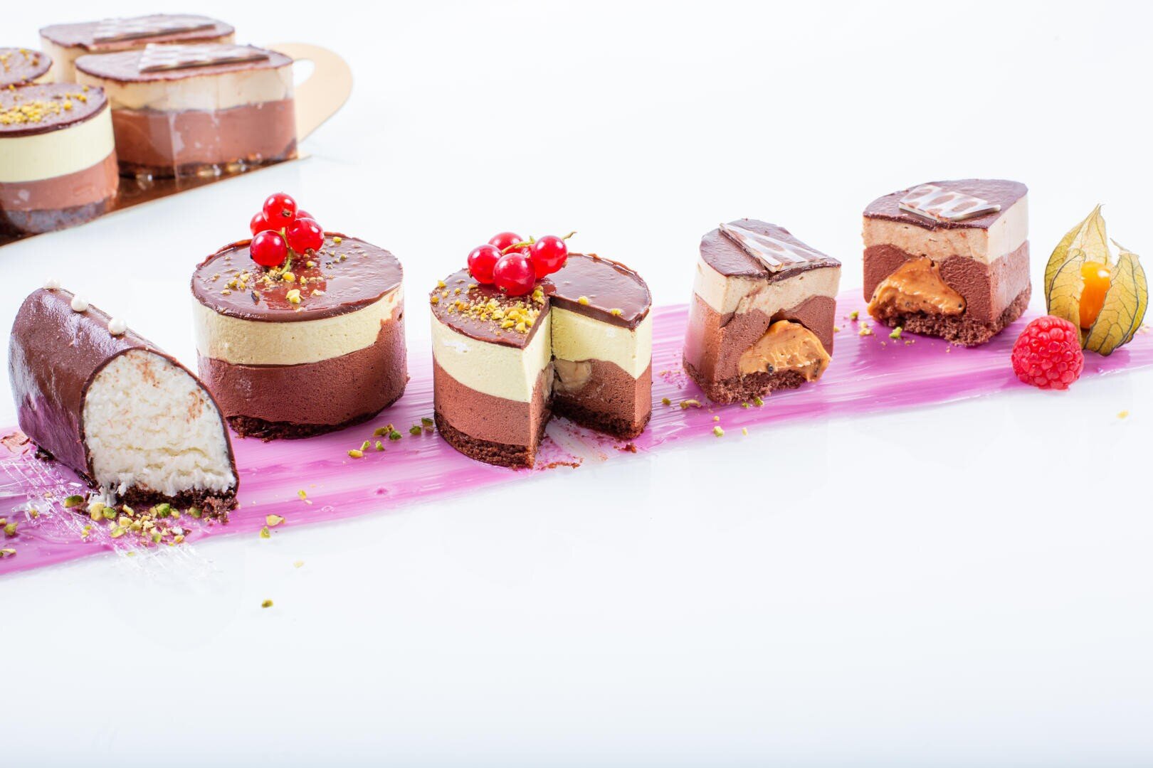 An assortment of chocolate mousse cakes and slices, some topped with red berries, are arranged on a pink streaked surface against a white background. A few cakes are cut open, revealing layered fillings.