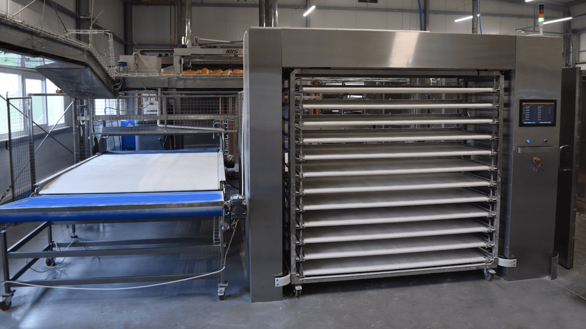 Industrial bakery equipment in a factory setting, featuring a multi-rack oven with a digital control panel and a conveyor belt system for automated bread or pastry production. Stainless steel surfaces and bright lighting are visible.