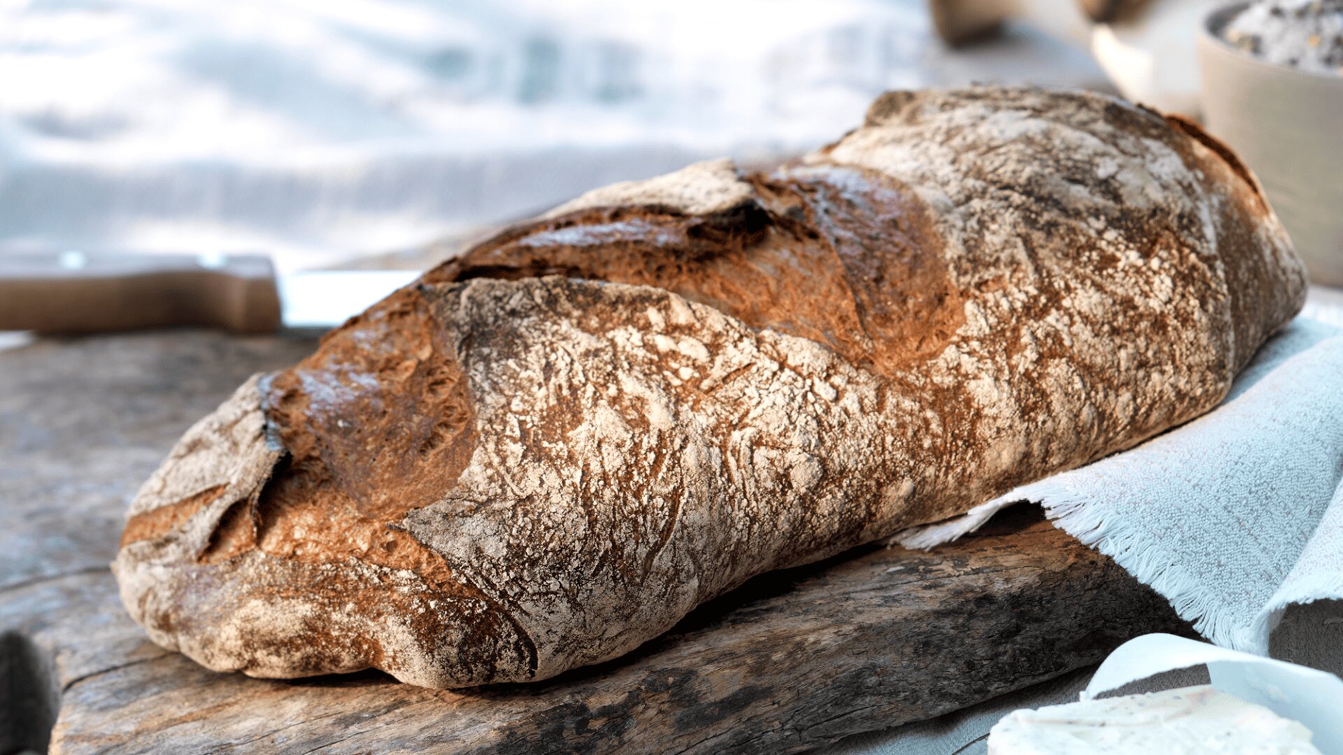 A rustic loaf of crusty artisan bread rests on a wooden board, next to a white cloth. The bread has a golden-brown, textured crust and an irregular shape, highlighted by natural light.