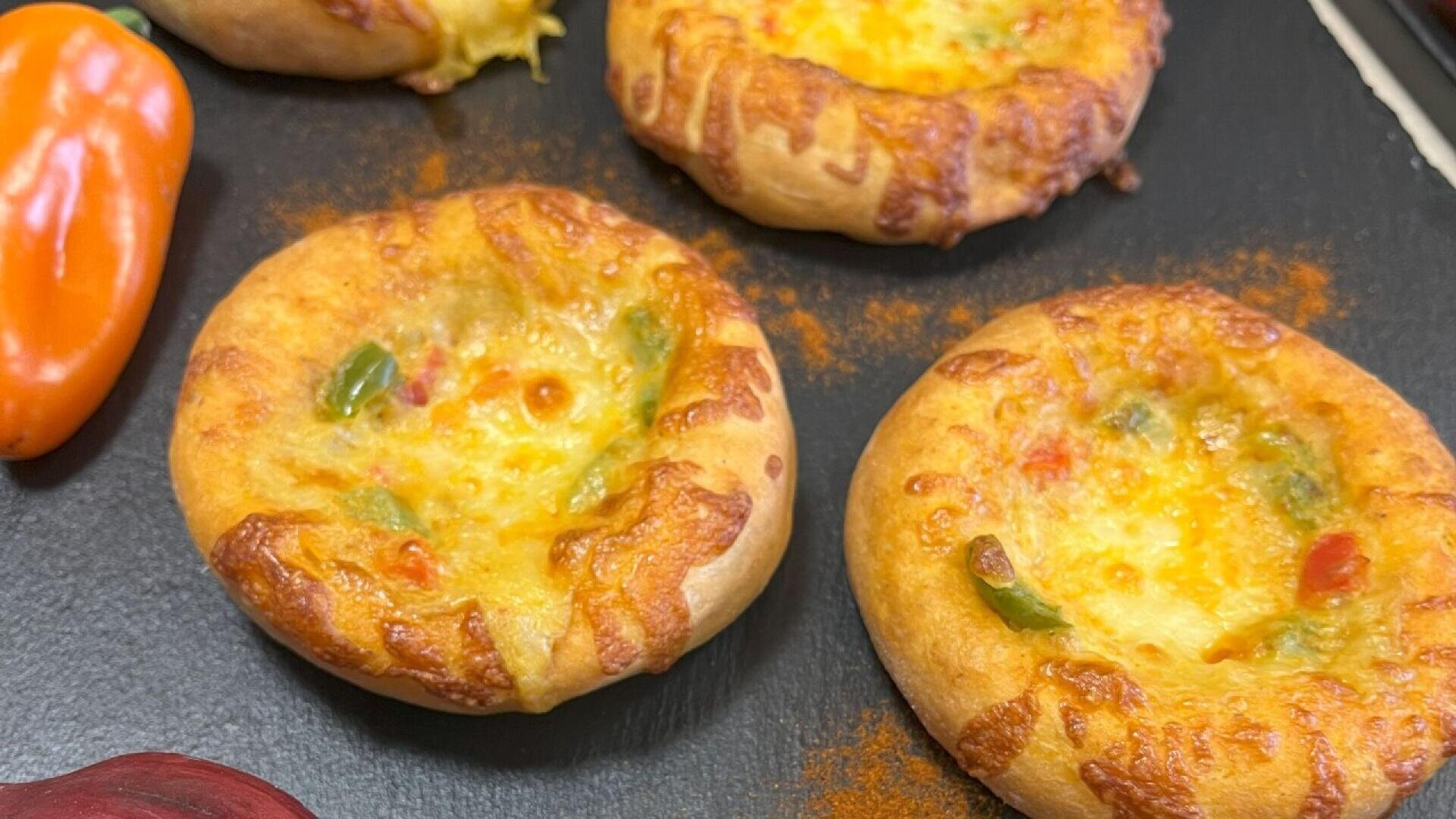 Four small, round bread rolls topped with melted cheese and colorful diced peppers sit on a black surface. A small orange pepper is visible on the left edge of the image.