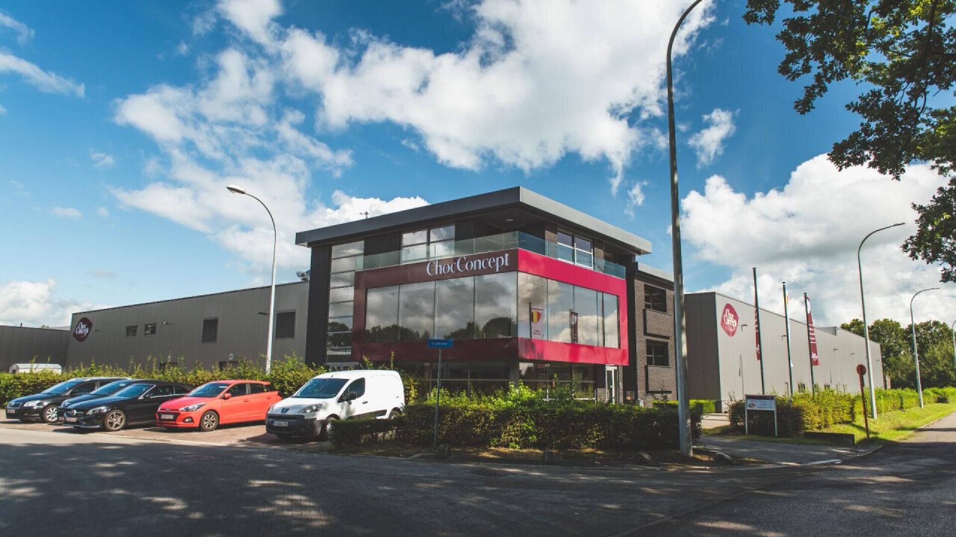 A modern commercial building with large windows and red accents, labeled Chox Concept. Several cars are parked outside under a partly cloudy sky, with trees and streetlights lining the street.