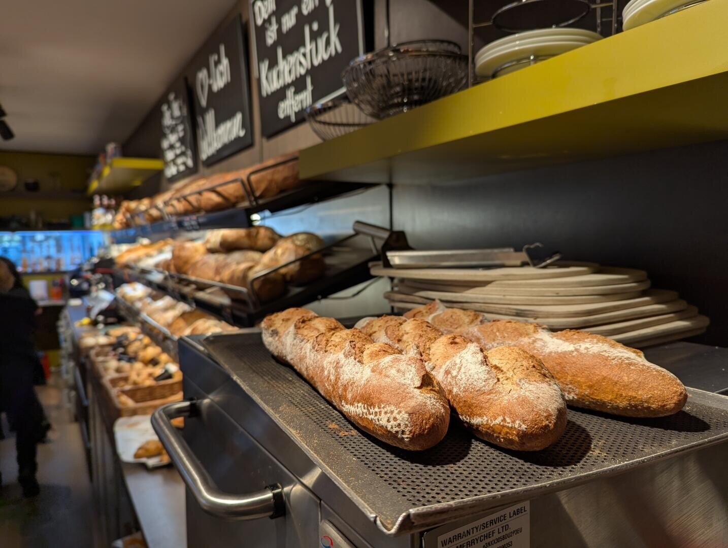 Frische Baguettes auf einem Metalltablett in einer Bäckerei mit Regalen voller Brot und Gebäck, darüber hängen Speisekarten auf Kreidetafeln, daneben sind Teller gestapelt. Der Laden ist warm beleuchtet.