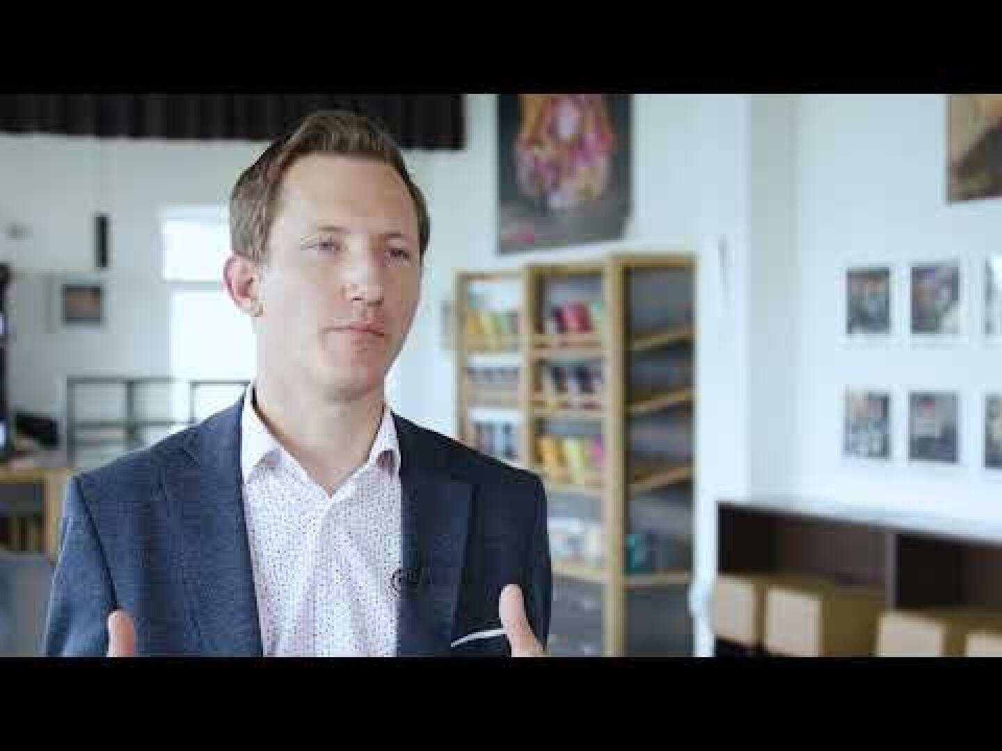 A man in a blazer and patterned shirt speaks while gesturing with his hands in a bright room with bookshelves, tables, and framed pictures on the walls in the background.