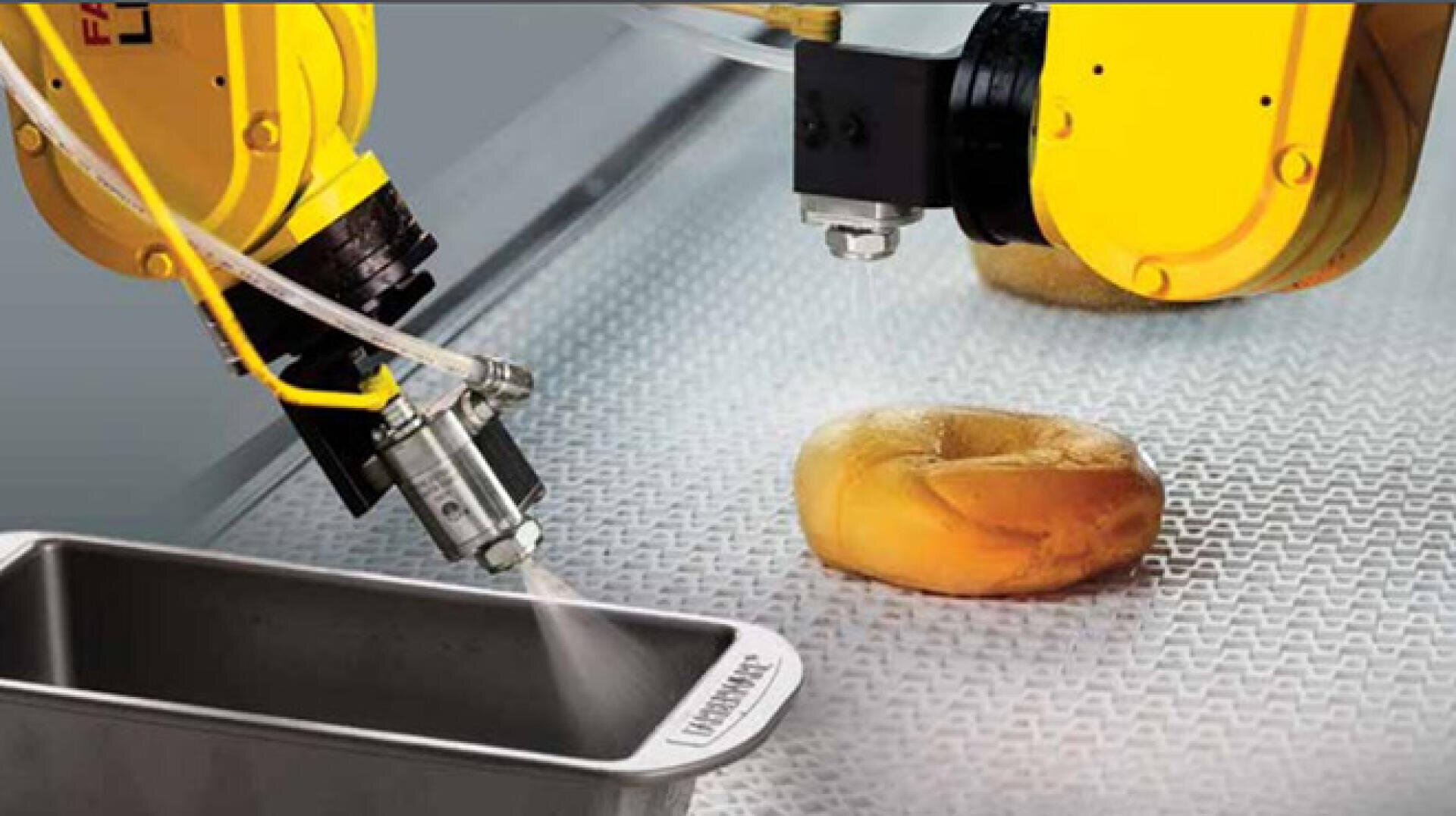 Ein Roboterarm sprüht Öl auf eine Brotbackform, während ein anderer Roboterarm eine Düse in die Nähe eines Bagels auf einem Förderband hält, was die automatisierte Lebensmittelverarbeitung veranschaulicht.