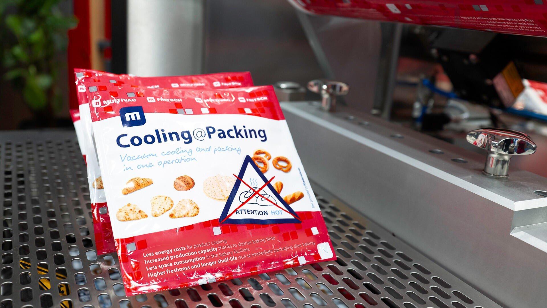 Zwei rot-weiße Pakete mit der Aufschrift „Cooling@Packing“ und Bildern von Backwaren stehen auf einer Metalloberfläche in der Industrie, neben Maschinen. Ein Schild auf dem Paket warnt: „Achtung: Heiß.“.