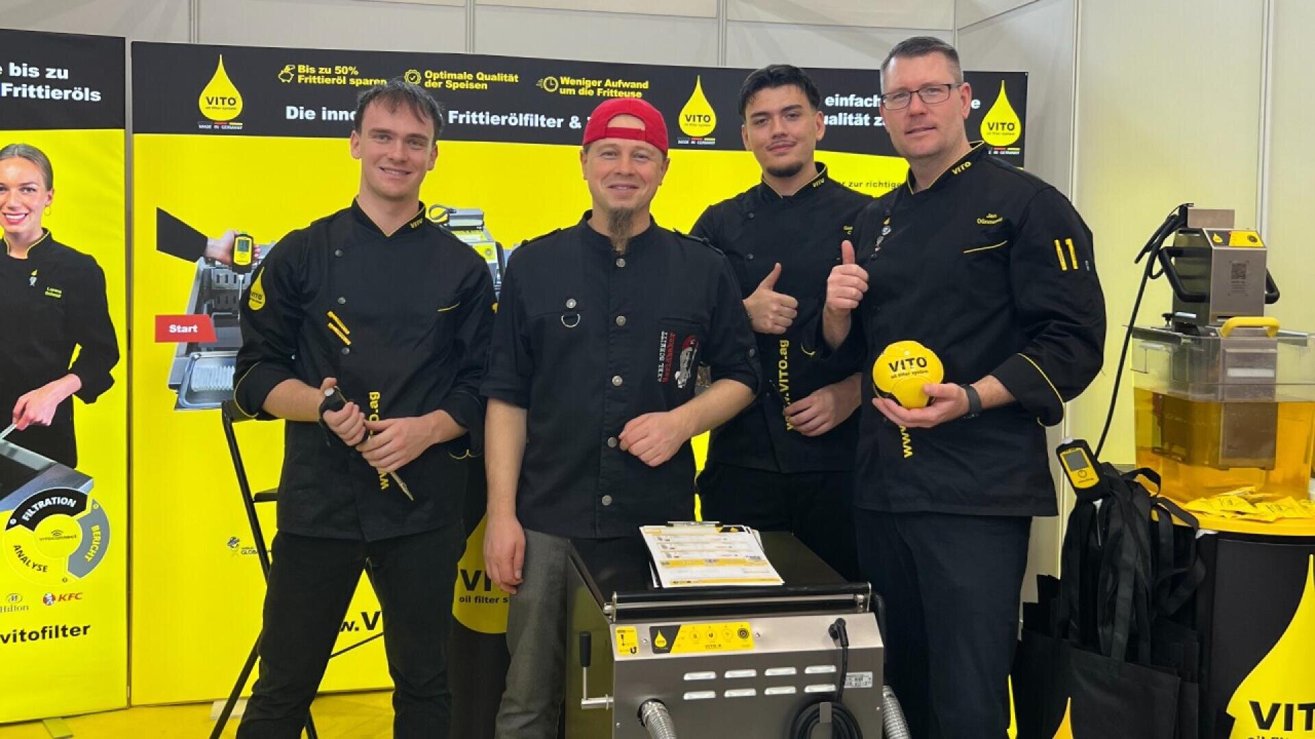 Four men in black uniforms stand smiling at a trade show booth with bright yellow Vito branding. One man wears a red hat, and another holds a yellow promotional item. They pose in front of a fryer and filter equipment.