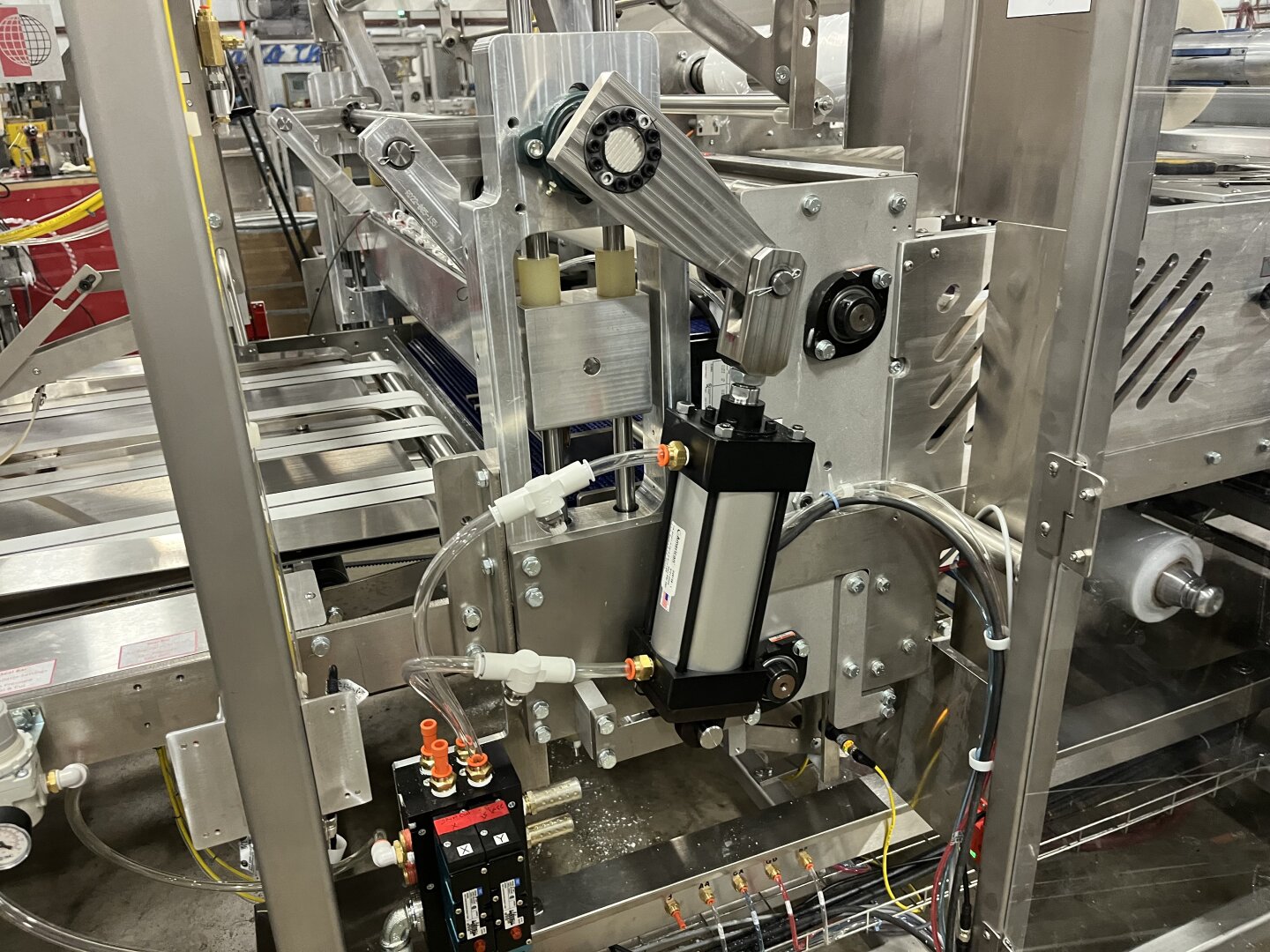 Nahaufnahme einer industriellen Verpackungsmaschine mit Metallrahmen, Rollen, Pneumatikzylindern und Drähten, die komplexe Montage- und Automatisierungsteile in einer Fabrikumgebung zeigt.