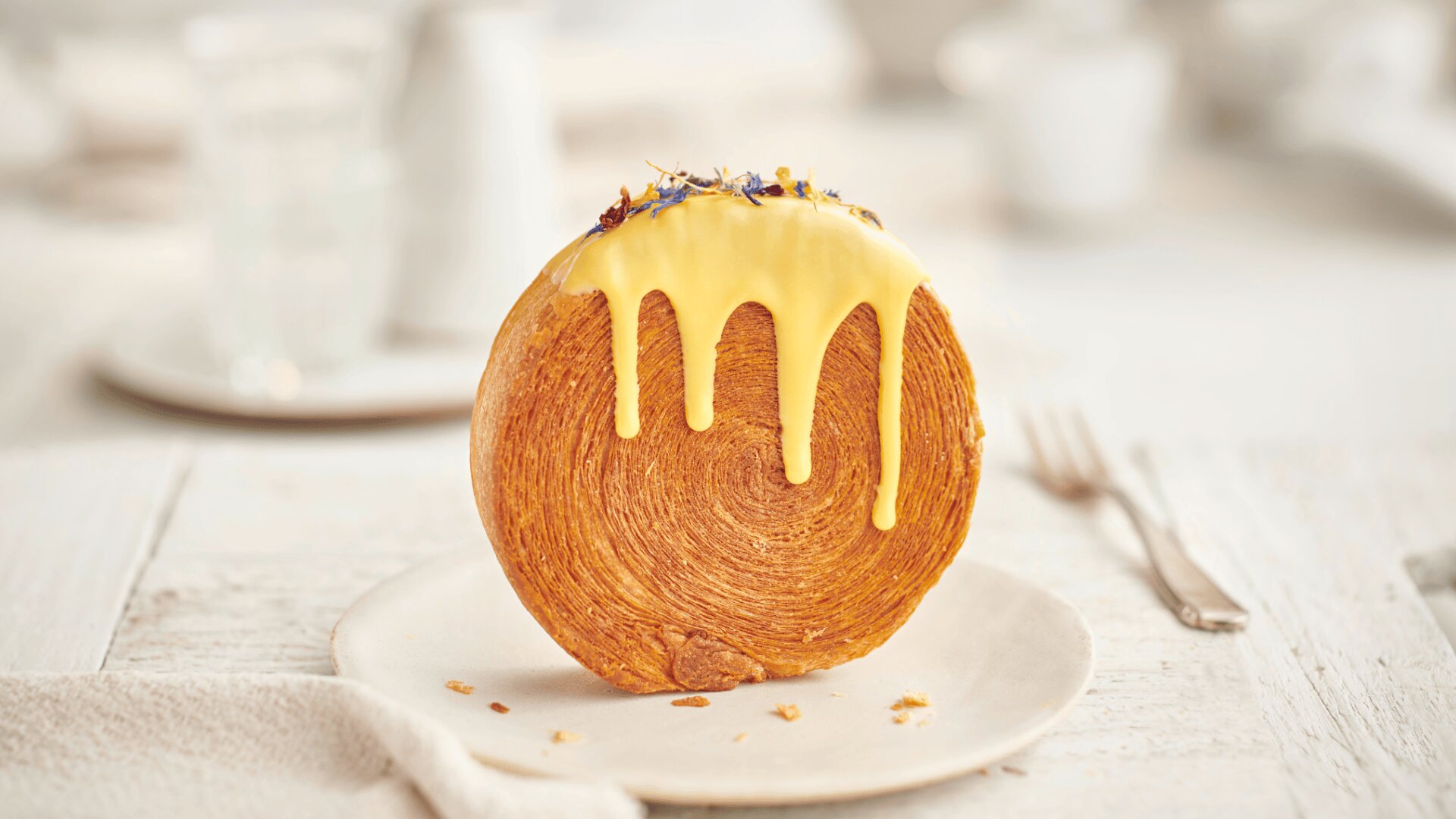 A round, flaky pastry stands upright on a white plate, topped with dripping yellow icing and small colorful edible flowers; a fork and blurred background objects are visible.