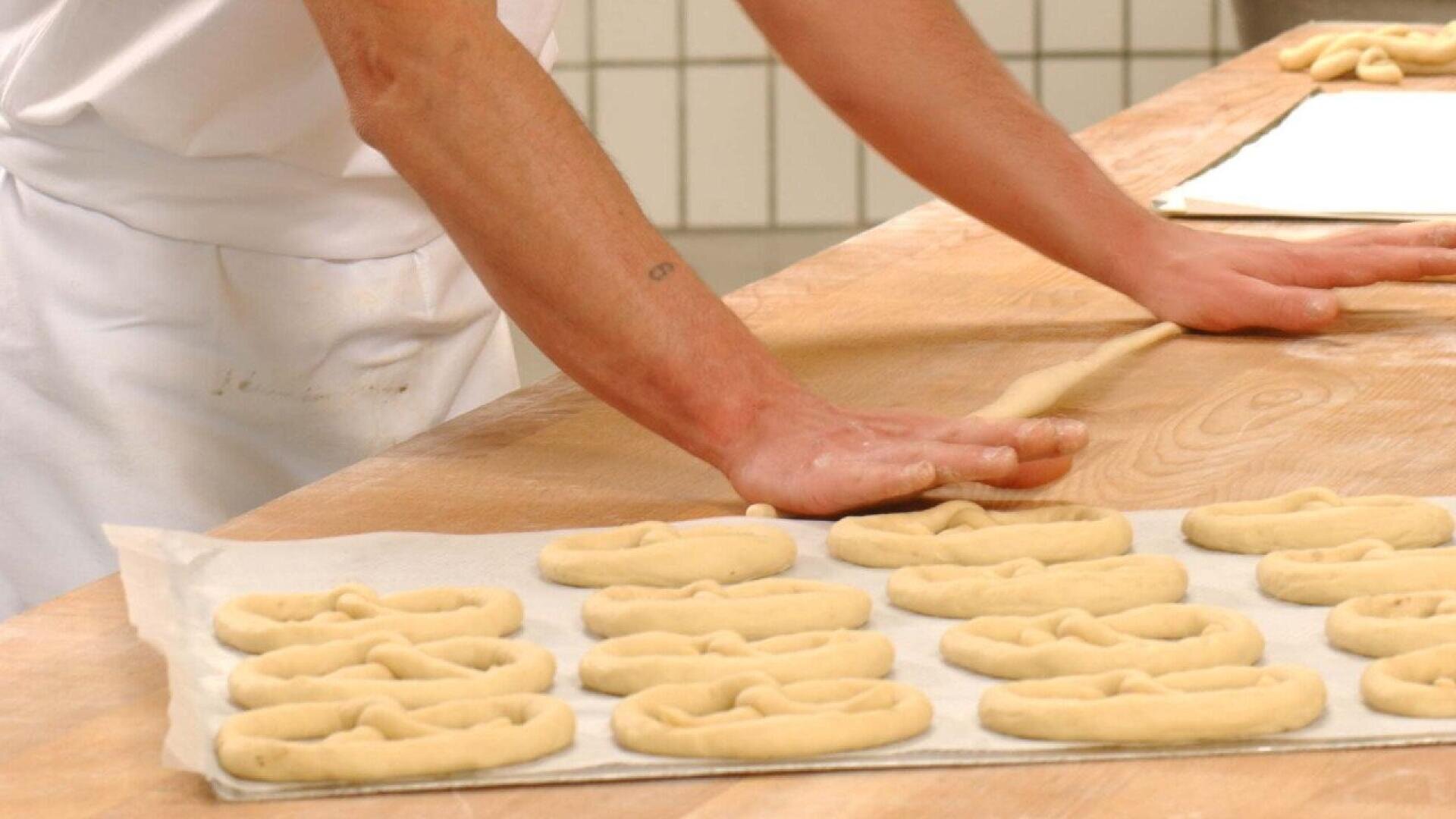 A person in a white apron rolls dough on a wooden surface, shaping it into pretzels. Several unbaked pretzels are arranged neatly on a parchment-lined baking tray nearby.