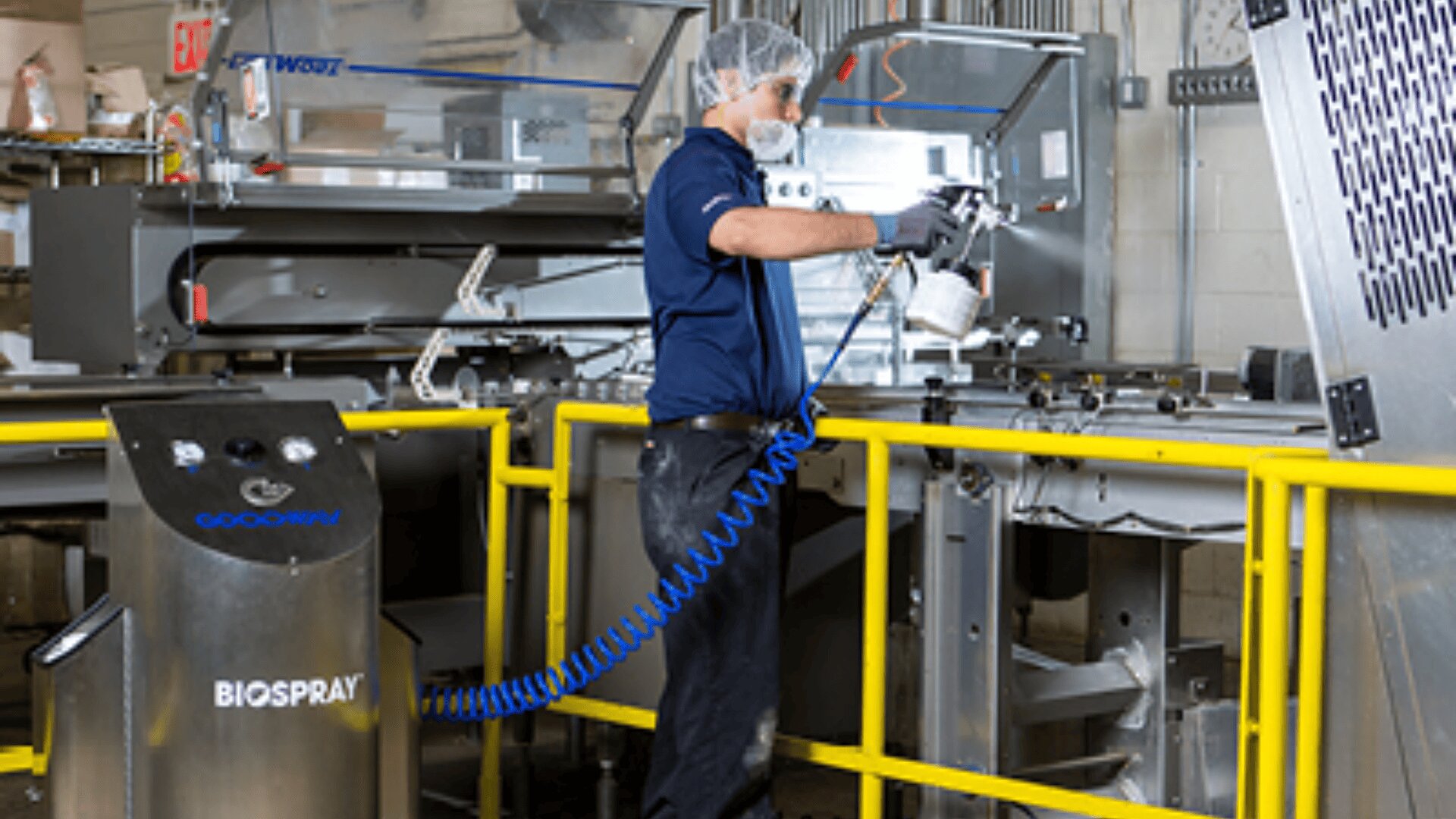 A worker wearing protective gear uses a BIOSPRAY machine to clean or sanitize industrial equipment in a factory setting, surrounded by metal machinery and safety railings.