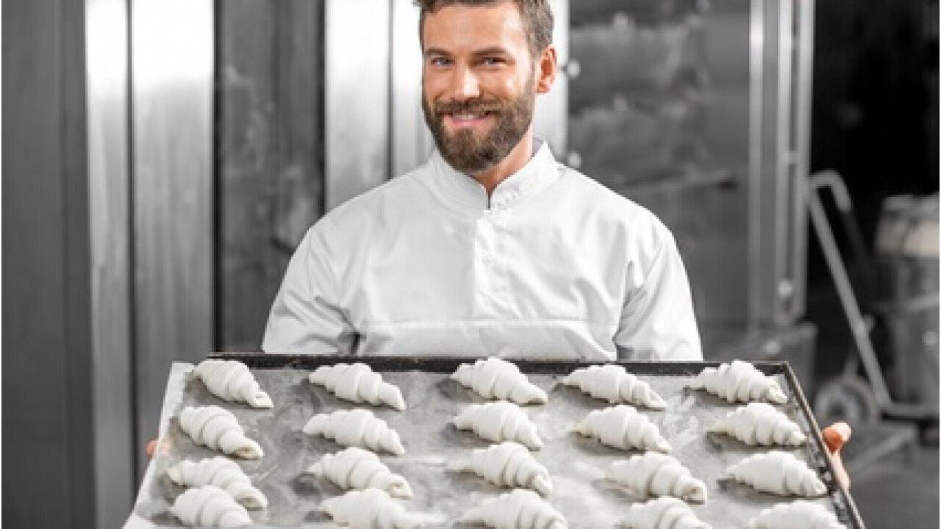A smiling baker in a white uniform holds a tray filled with rows of unbaked croissants in a bakery kitchen.