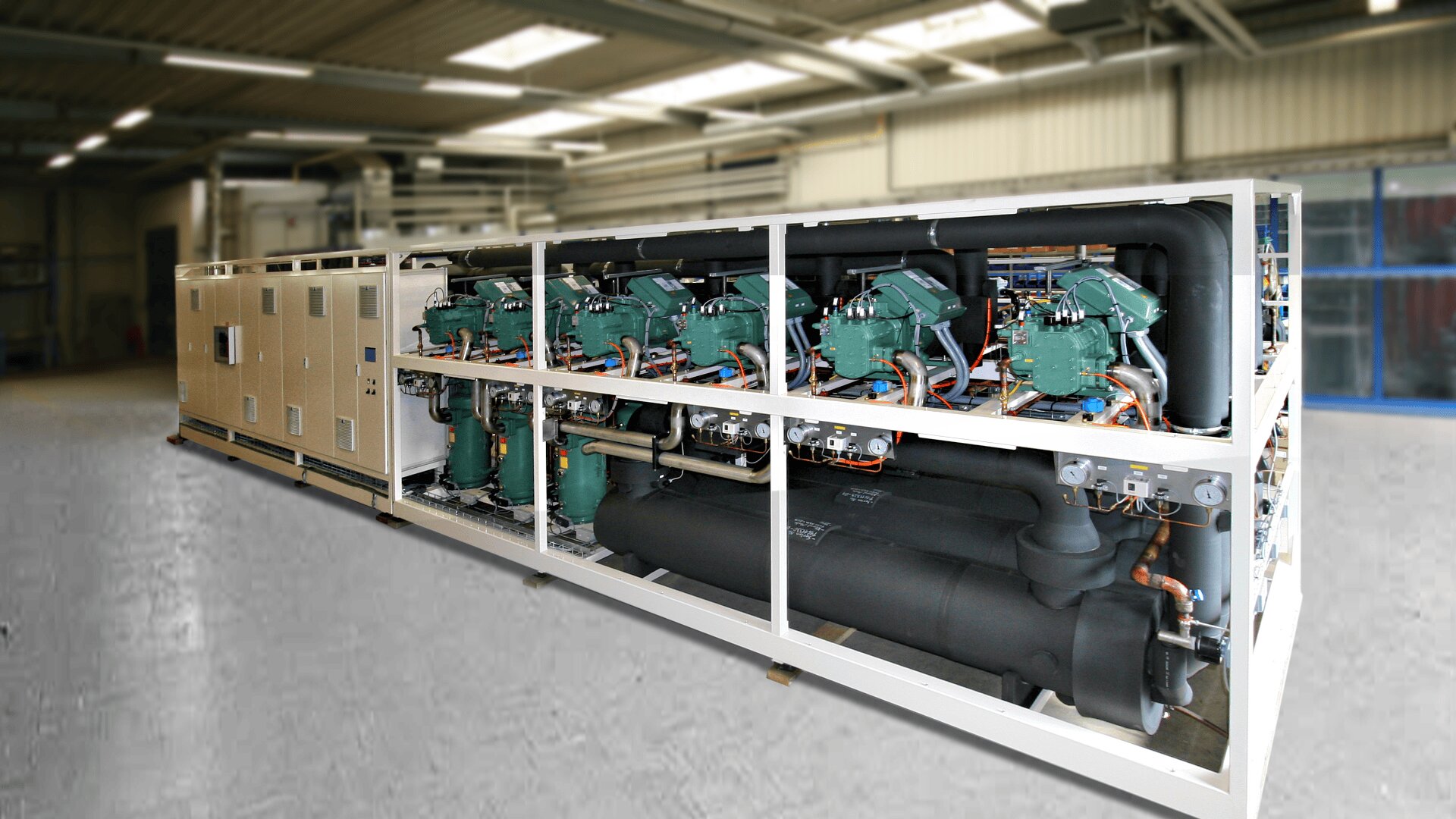 Ein großes industrielles Kühlaggregat mit mehreren Verdichtern und Rohrleitungen, das in einem Metallrahmen untergebracht ist und in einem Innenraum auf einem Betonboden steht.
