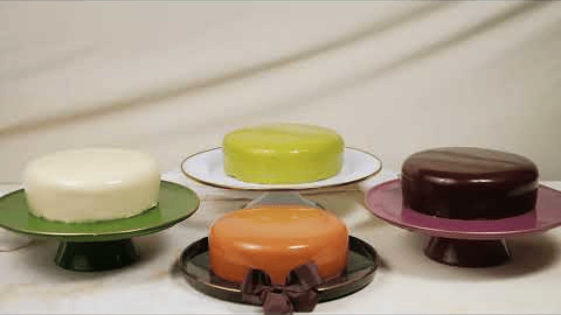 Four glossy, round cakes in different colors—white, light green, orange with a brown ribbon, and dark brown—sit on various cake stands against a neutral draped background.