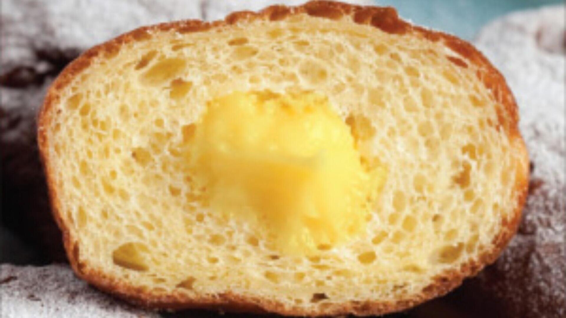 Eine Nahaufnahme eines halbierten Teigs, der ein fluffiges, luftiges Inneres mit einer cremigen gelben Füllung in der Mitte zeigt. Die äußere Kruste ist goldbraun und leicht knusprig.