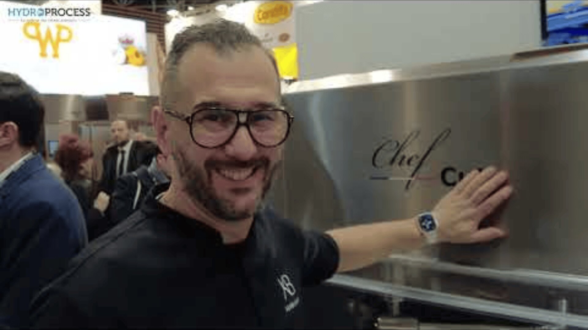 A smiling man with glasses and a beard stands next to a large metal appliance labeled Chef, resting his hand on it. He is indoors at an event with people and booths in the background.