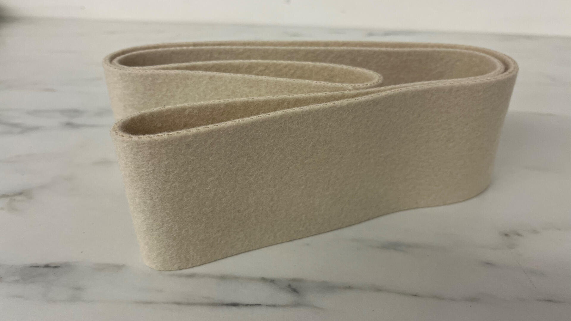 Ein beiger Filzstreifen ist in einer losen, akkordeonartigen Form auf einer weißen Marmorfläche gefaltet.