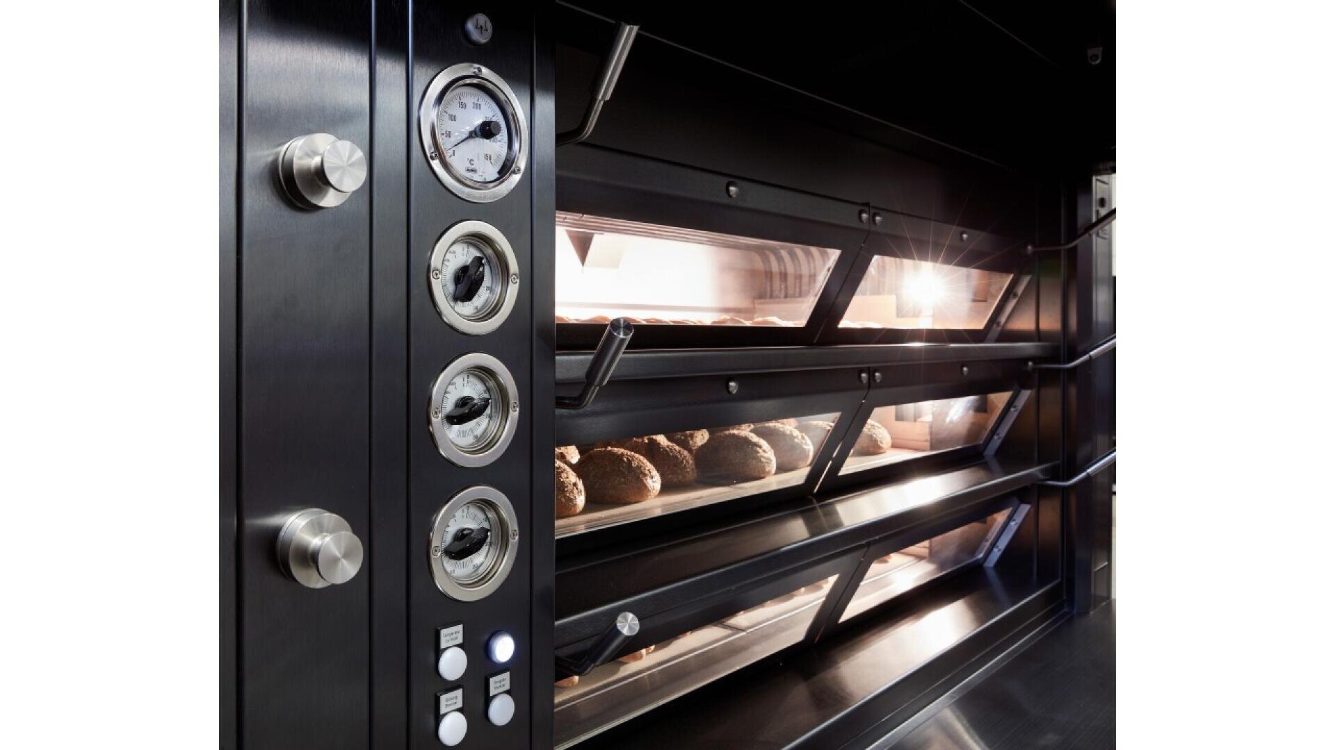 Close-up of a commercial bakery oven with three shelves, each containing loaves of bread baking. The oven features dials, gauges, and buttons on the side, and the interior is brightly lit.