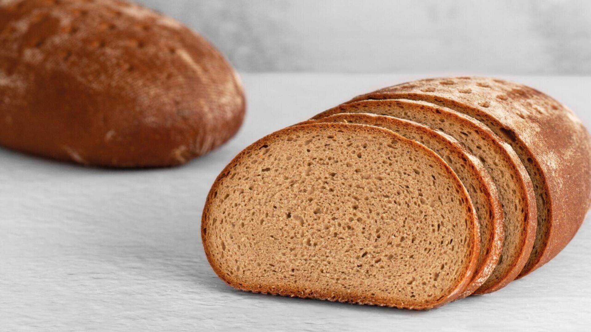 A loaf of brown bread with several slices cut and arranged in front of the unsliced portion, resting on a light-colored surface with another whole loaf in the background.