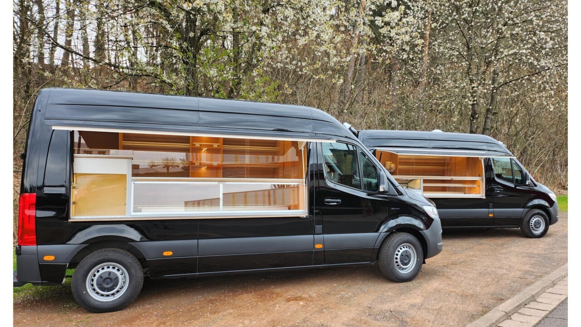 Two black vans with large side windows display modern, well-lit wooden interiors, parked outdoors on a dirt road with blooming trees in the background.