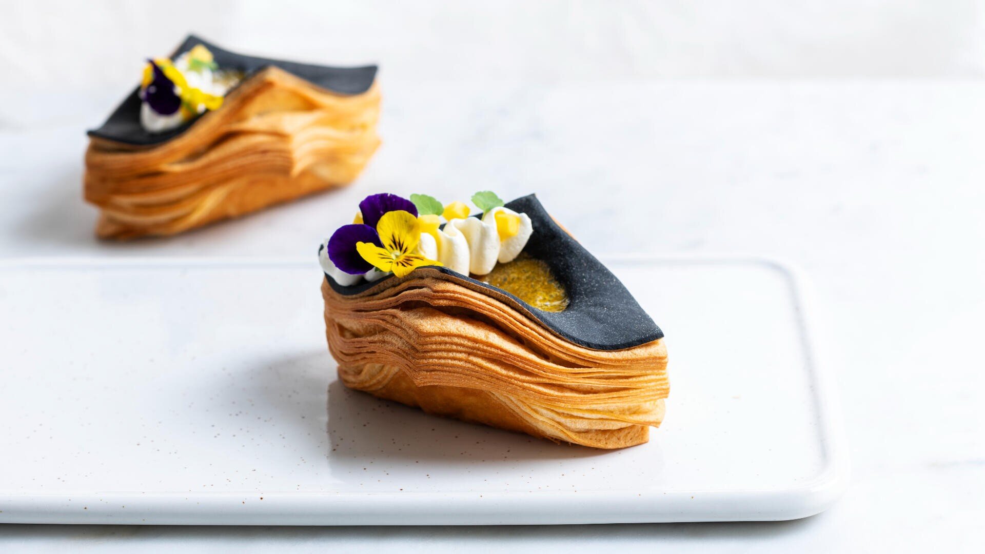 A stylish pastry with flaky golden layers, topped with black icing, cream, and edible purple, yellow, and white flowers, sits on a white rectangular plate with another similar pastry in the background.