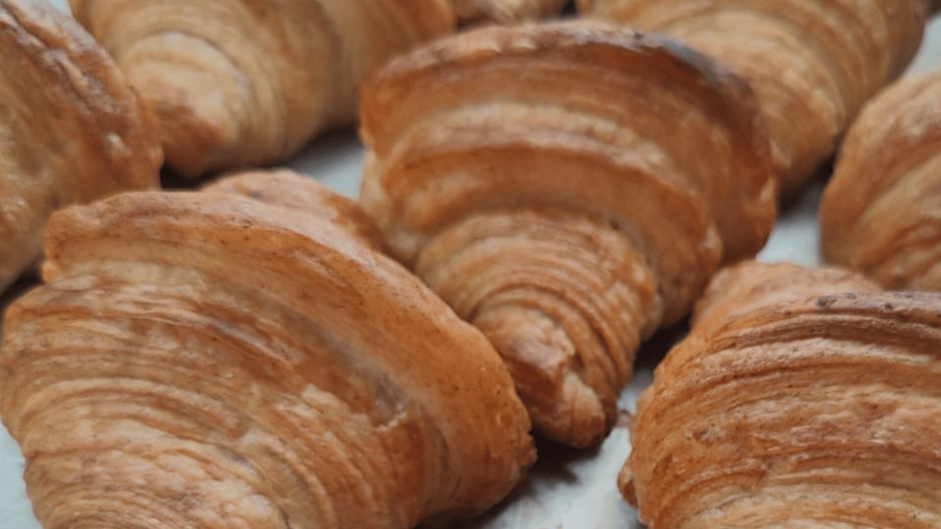 A close-up of several golden brown croissants with flaky, layered pastry, arranged on a light surface.