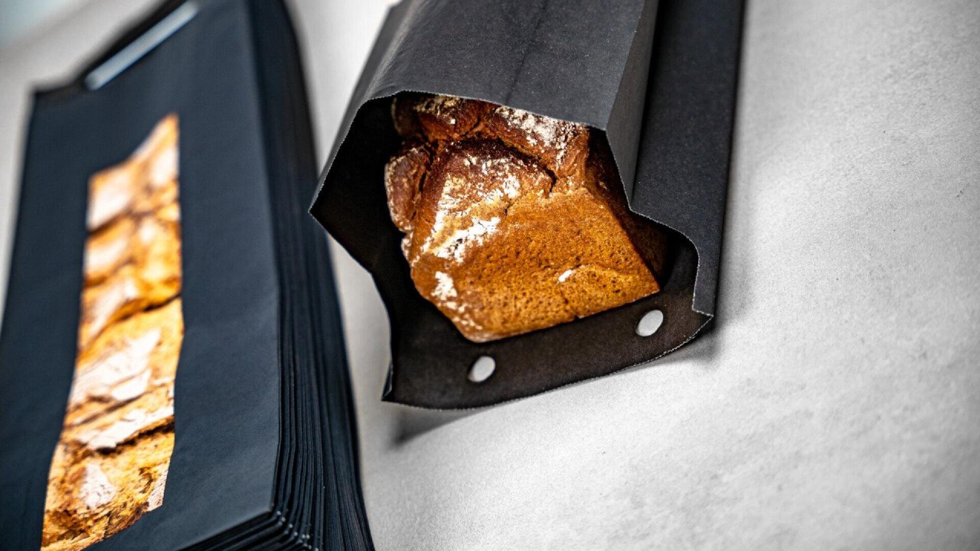 A shiny loaf of bread is partially wrapped in a black paper bag on a light surface, with a stack of similar black bags and baked goods visible nearby.