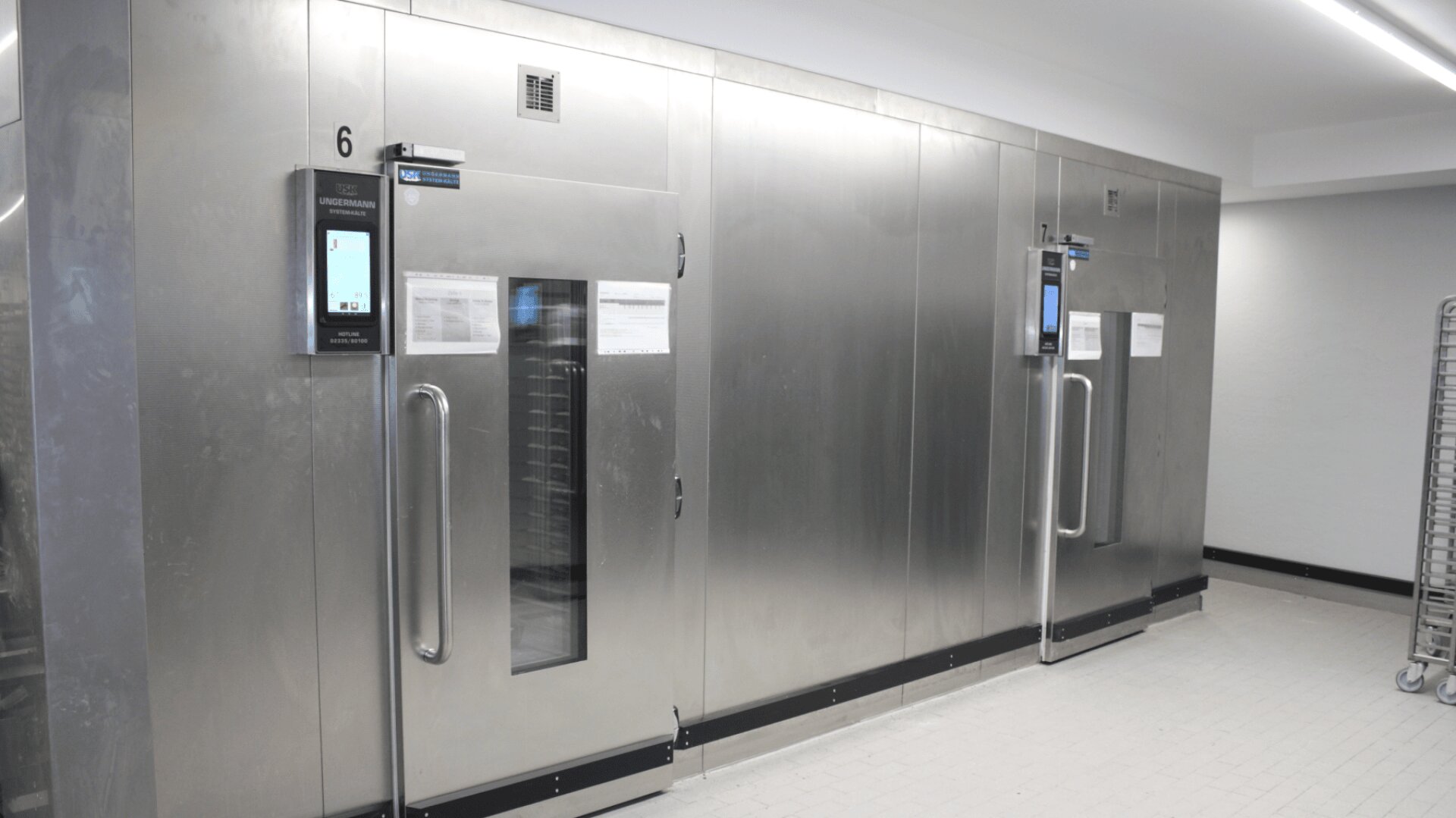 Drei große Industriekühlschränke aus Edelstahl mit Glastüren und elektronischen Schalttafeln stehen nebeneinander in einem sauberen, weiß gefliesten Raum.