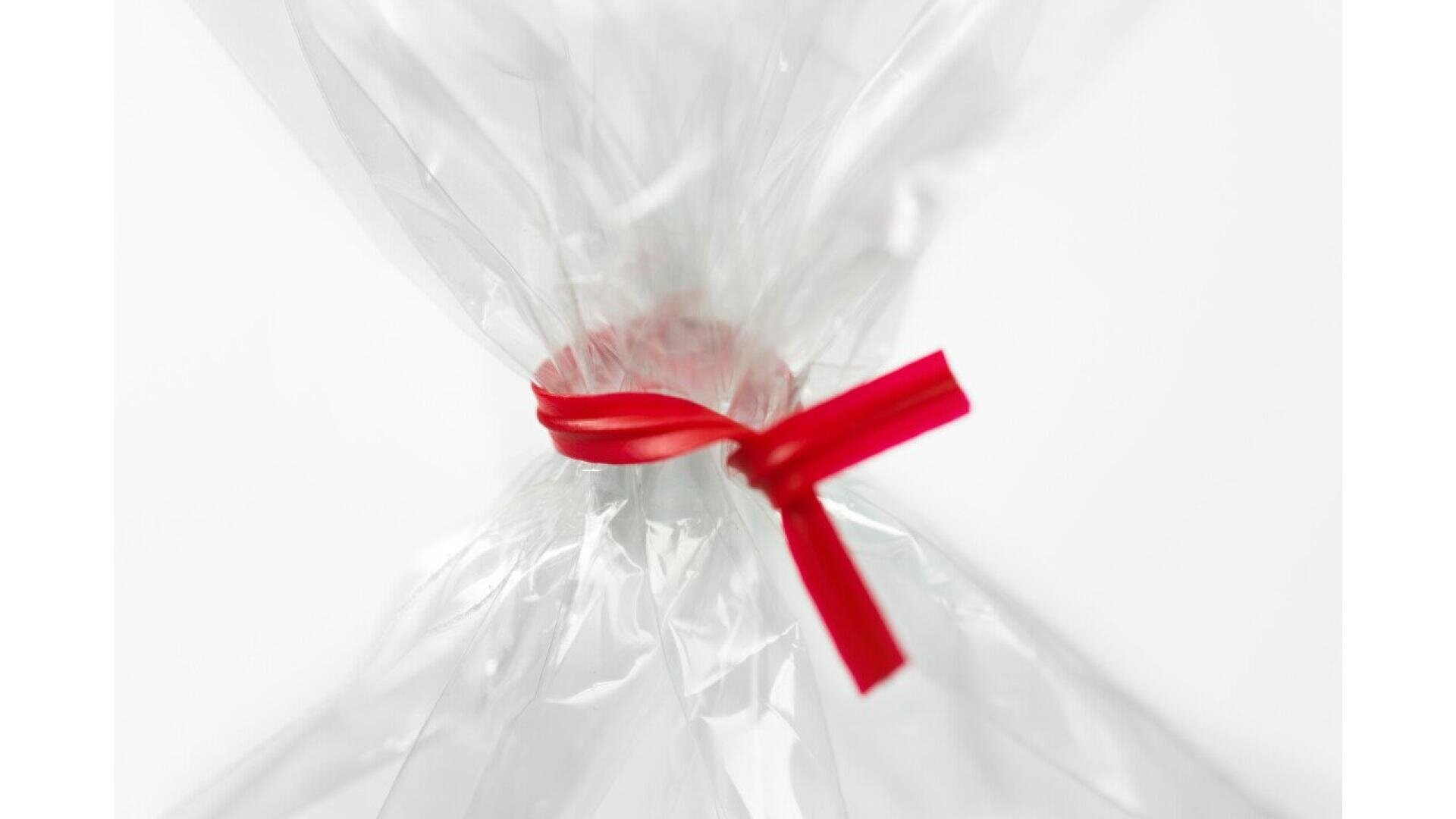 Transparente Zellophanverpackung, oben mit einem roten Band vor weißem Hintergrund verschnürt. Der Schwerpunkt liegt auf dem Knoten, wobei das Plastik fest zusammengerafft ist und die Enden sich oben und unten auffächern.
