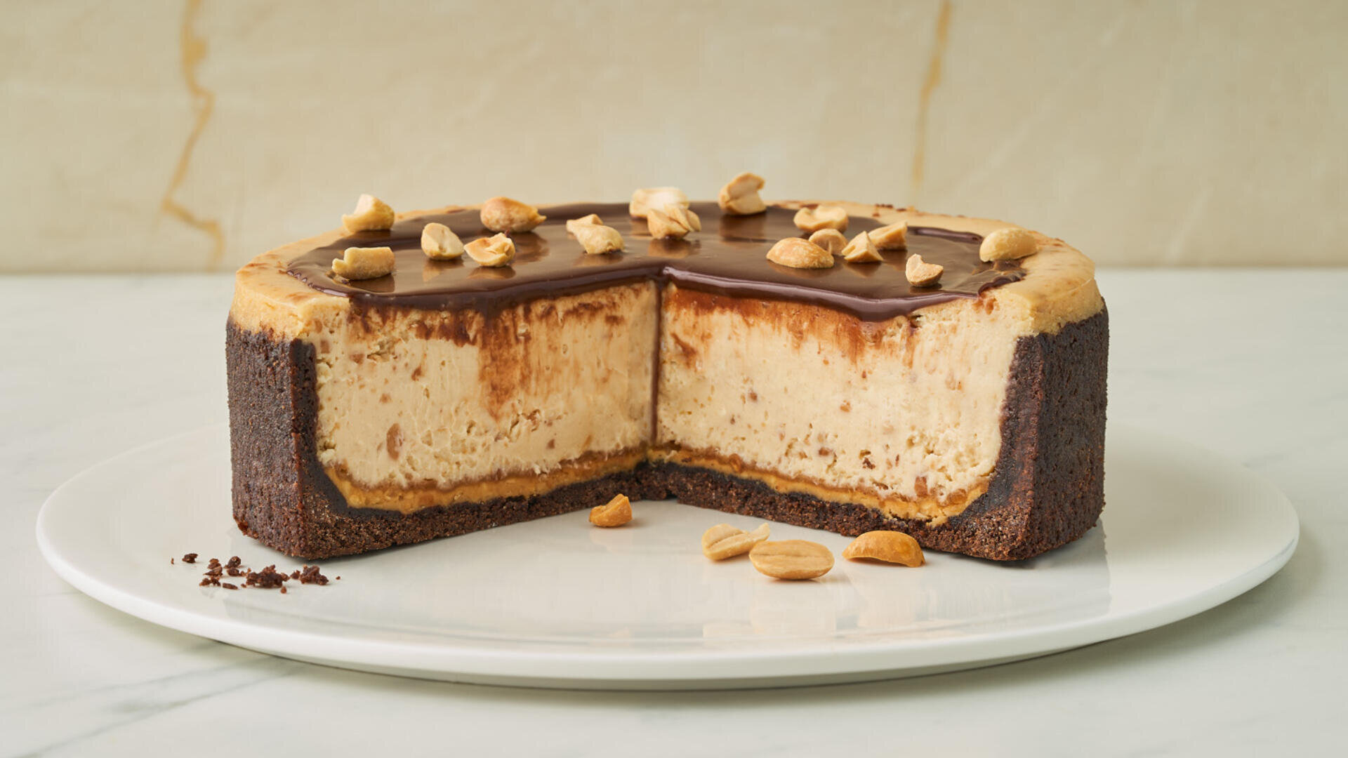 A round cheesecake with a chocolate crust and chocolate topping, garnished with peanuts. A large slice has been removed, revealing the creamy filling inside. The cake sits on a white plate against a light marble background.