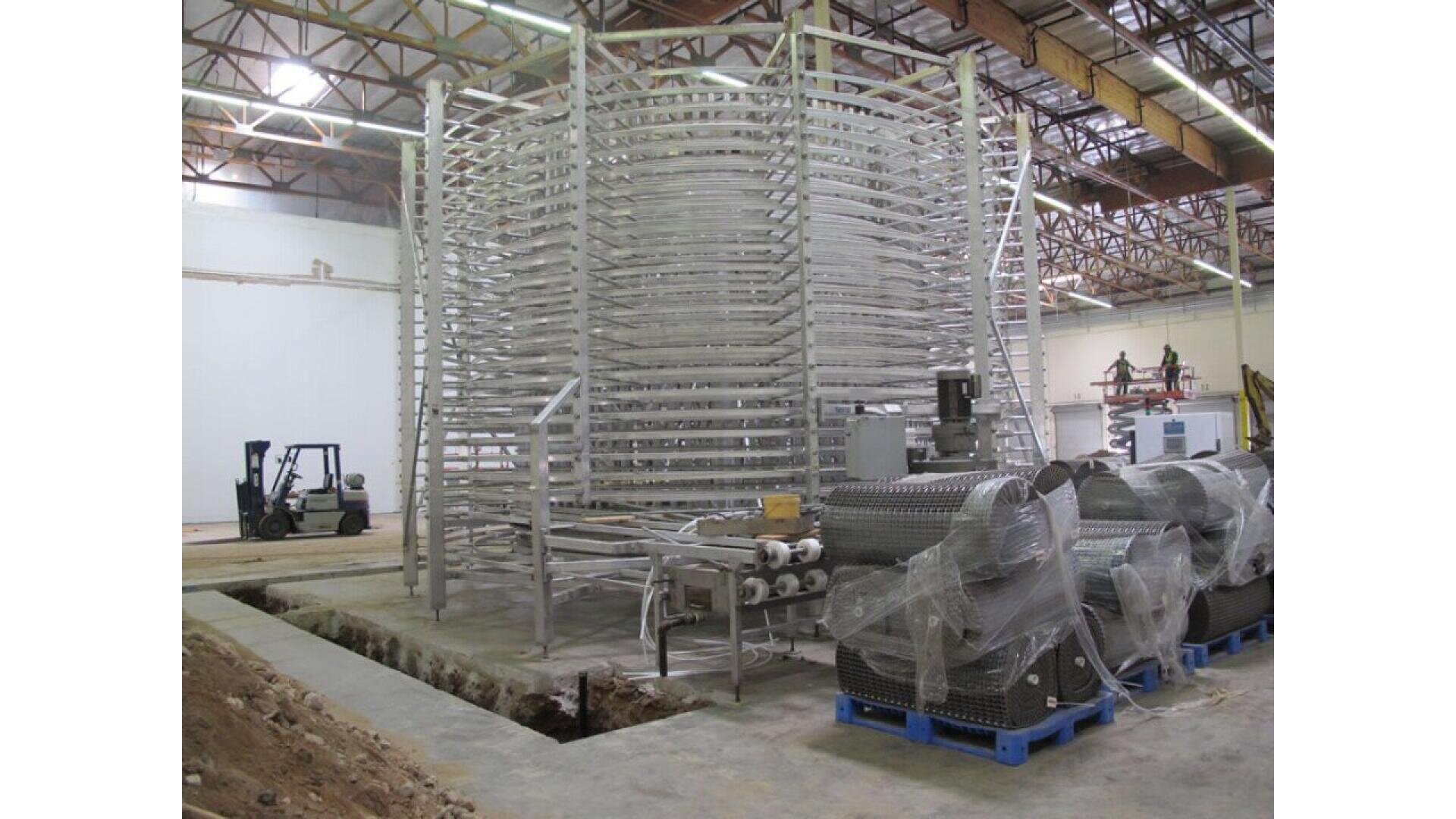 Großer industrieller Spiralgefrierschrank in einer Lagerhalle mit freiliegenden Deckenbalken, umgeben von Maschinen und einem Gabelstapler im Hintergrund; einige Geräte sind in Plastik verpackt und stehen auf blauen Paletten.