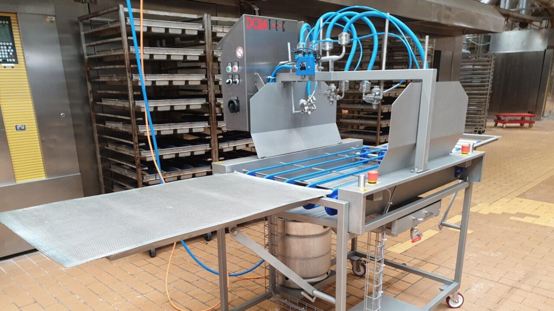 A stainless steel industrial machine with blue hoses and a conveyor, designed for food processing, stands in a commercial bakery with racks and ovens in the background.