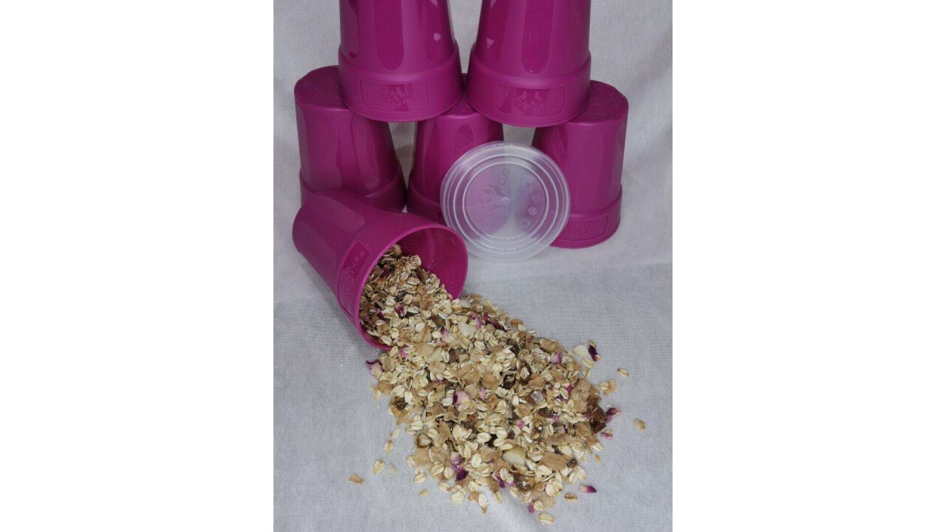 A stack of pink plastic cups, with one tipped over and spilling a mixture of oats, seeds, and dried flower petals onto a white surface. A clear plastic lid is also visible among the cups.