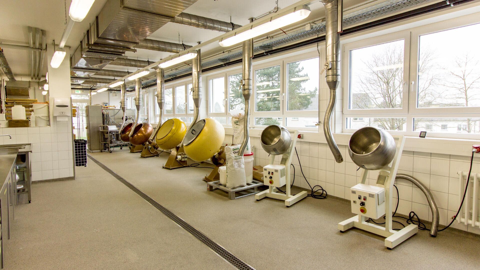 Eine helle Industrieküche mit großen Fenstern, mehreren Drumcomputern an der Wand, Lüftungsrohren darüber sowie gefliesten Wänden und Böden.