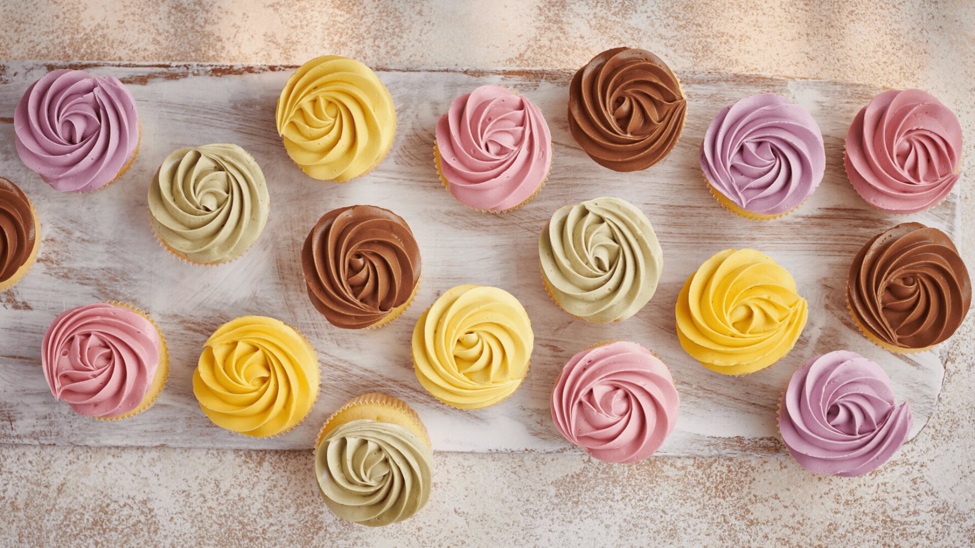 Verschiedene Cupcakes mit gewirbeltem Zuckerguss in Gelb, Rosa, Braun und Hellgrün sind auf einer weiß gekalkten Holzfläche angeordnet und bilden eine farbenfrohe und einladende Auslage.