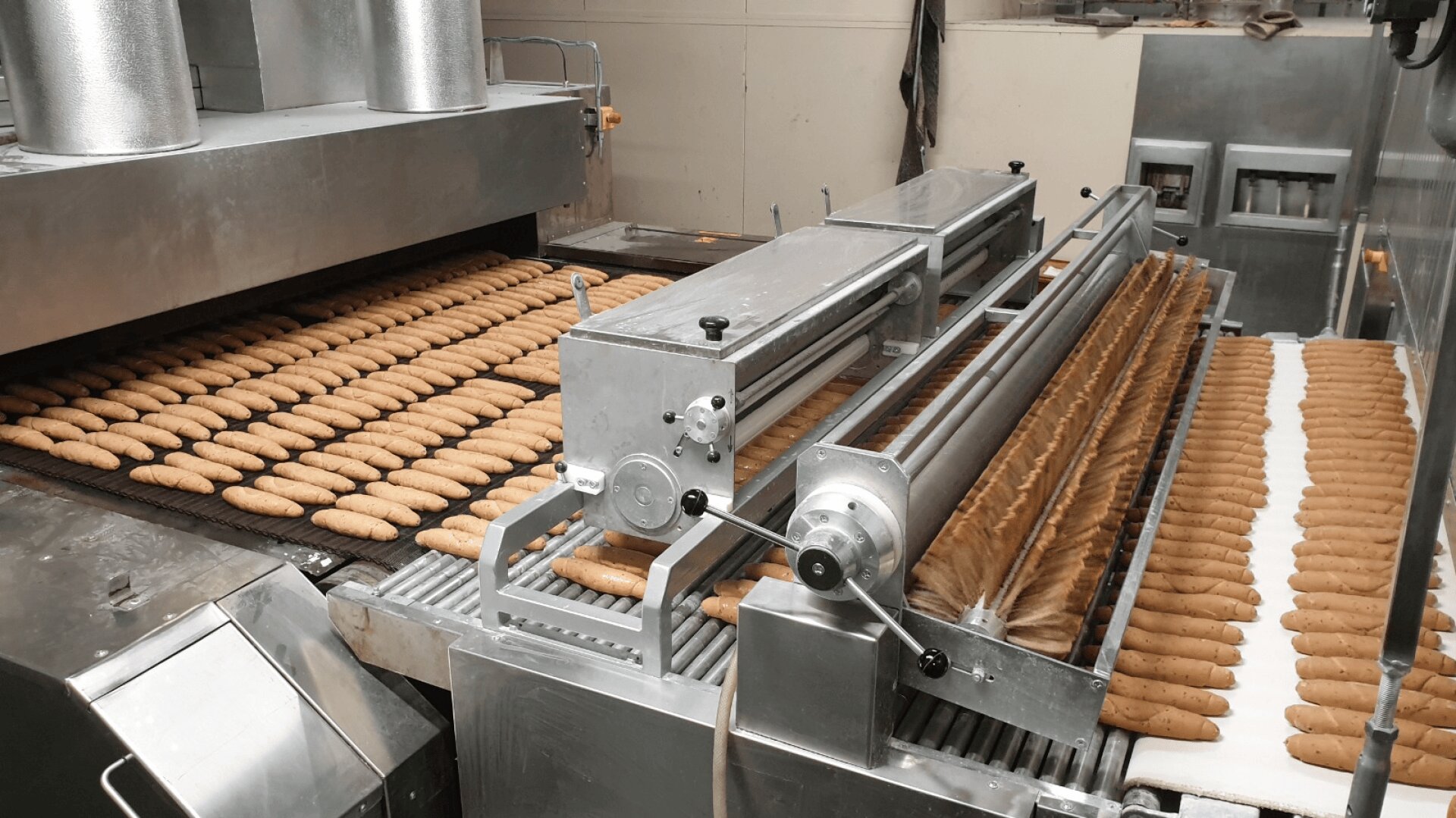 Rows of brown breadsticks move along an automated conveyor system in a bakery factory, passing through metal machinery for processing. The setting is industrial, with stainless steel equipment and breadsticks in uniform rows.