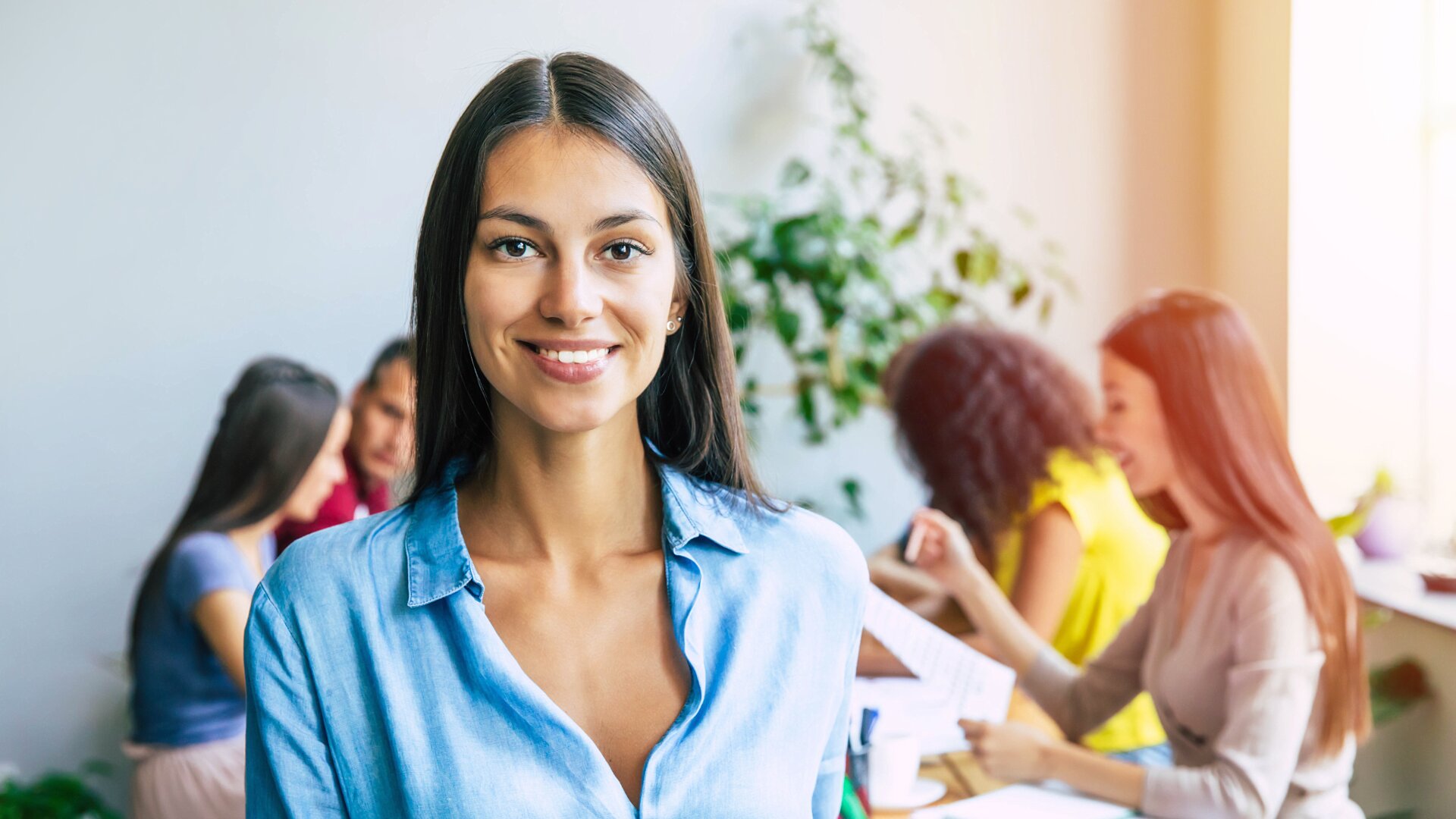 Eine Frau im blauen Hemd lächelt in die Kamera, während im Hintergrund drei Frauen an einem Tisch in einem hellen, modernen Büroraum mit Pflanzen arbeiten und sich unterhalten.