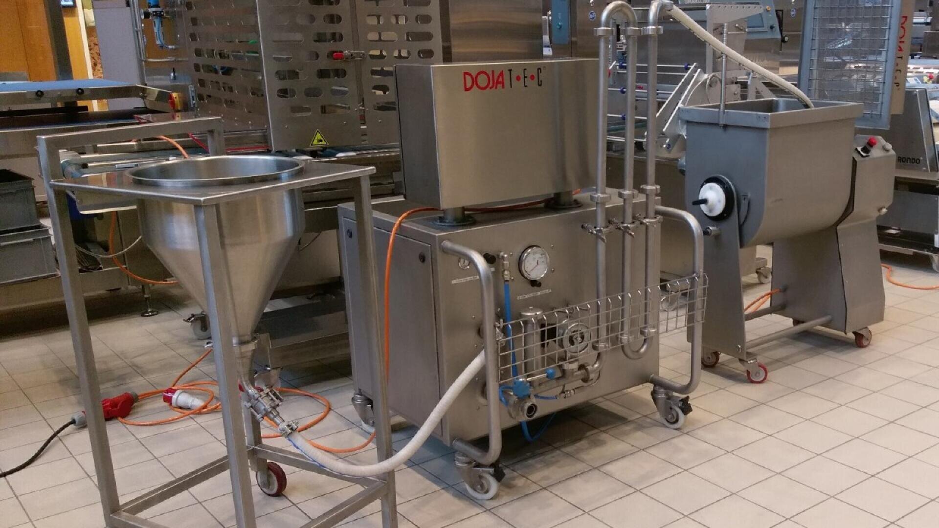 Industrial food processing equipment with stainless steel tanks, pipes, and gauges connected by hoses, standing on a tiled floor in a commercial kitchen or factory setting.