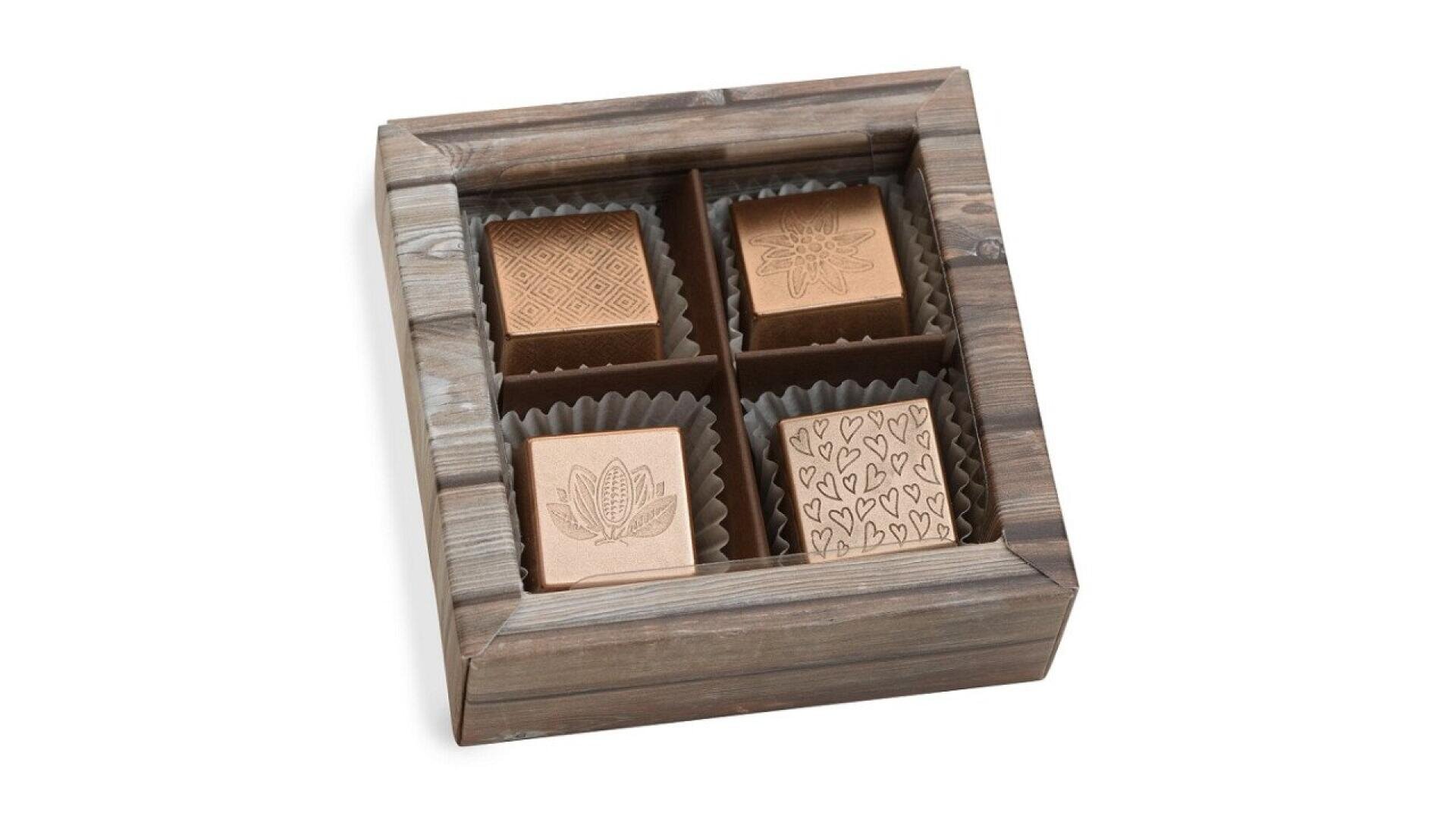 A wooden box containing four square chocolates, each decorated with a different engraved pattern, separated by dividers and placed in brown paper cups.