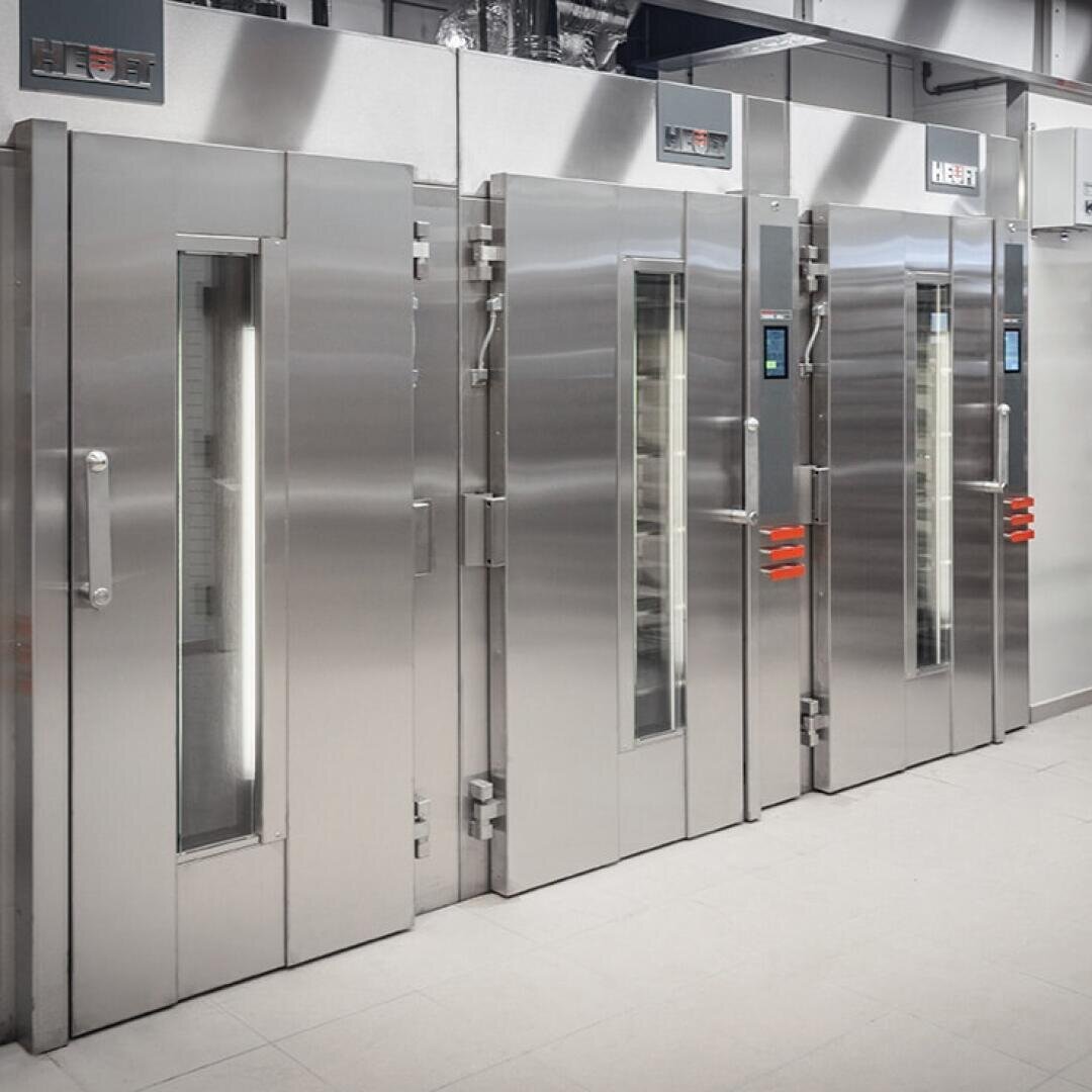 Drei große Industrieöfen aus Edelstahl mit vertikalen Fenstern und roten Griffen sind in einer sauberen, modernen Großküche nebeneinander aufgereiht.